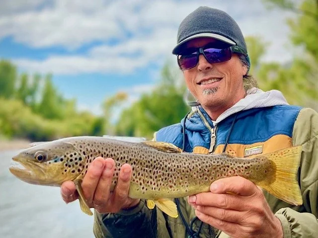 Man holding a large trout fish outdoors near a river, wearing sunglasses, a cap, and a blue jacket.