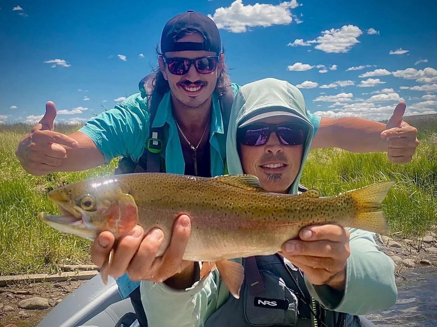 Two men outdoors, one holding a large fish, both wearing sunglasses and casual outdoor clothing, smiling. The background features green grass, a blue sky with clouds.
