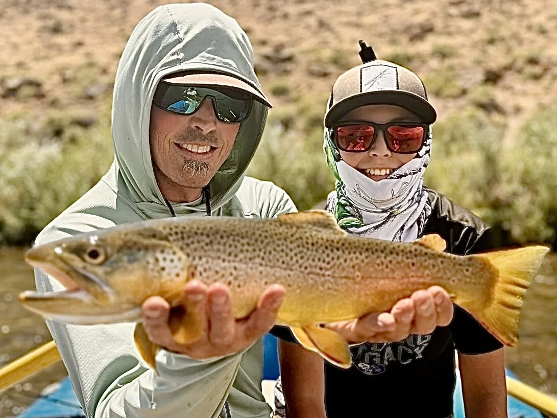 Two people smiling and holding a large rainbow trout fish outdoors near water, wearing sunglasses and hats.