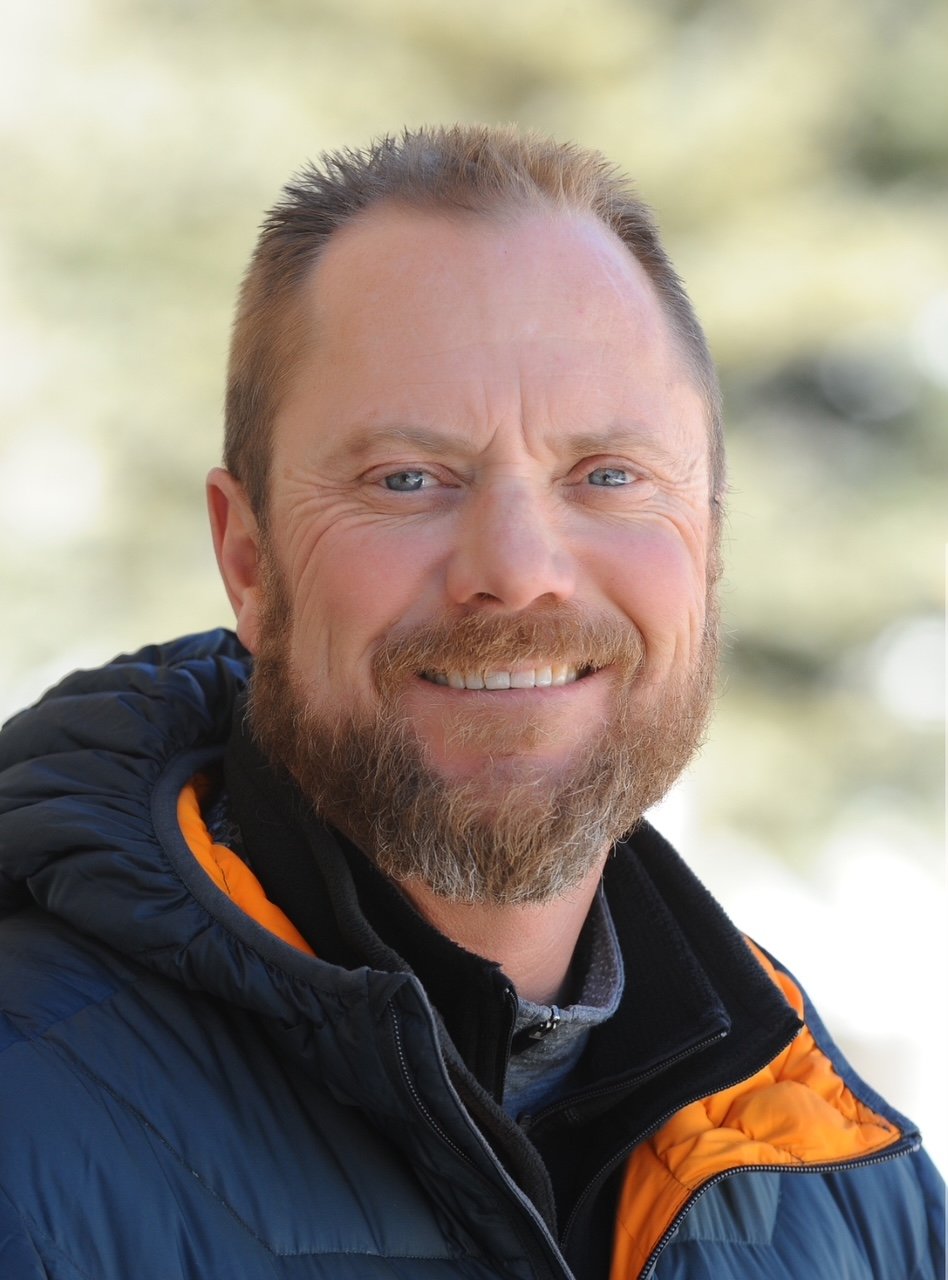 A smiling man with a beard and short hair, wearing a black and orange jacket, outdoors with blurred trees in the background.