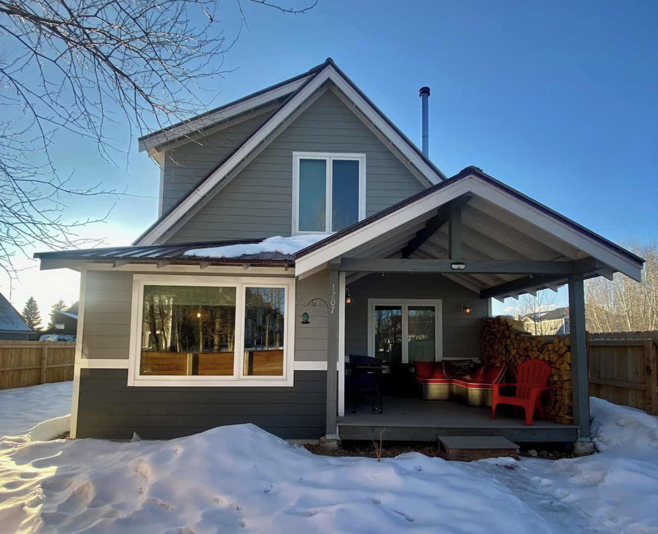 A two-story house with gray siding, a front porch with snow on the roof, and a stack of firewood on the porch, during winter daytime.