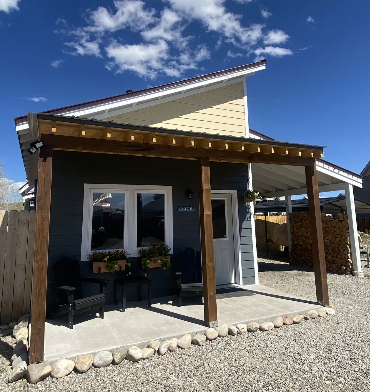 Front view of a modern house with a porch, wooden support beams, potted plants, a window, and a door. Blue sky with scattered clouds overhead.