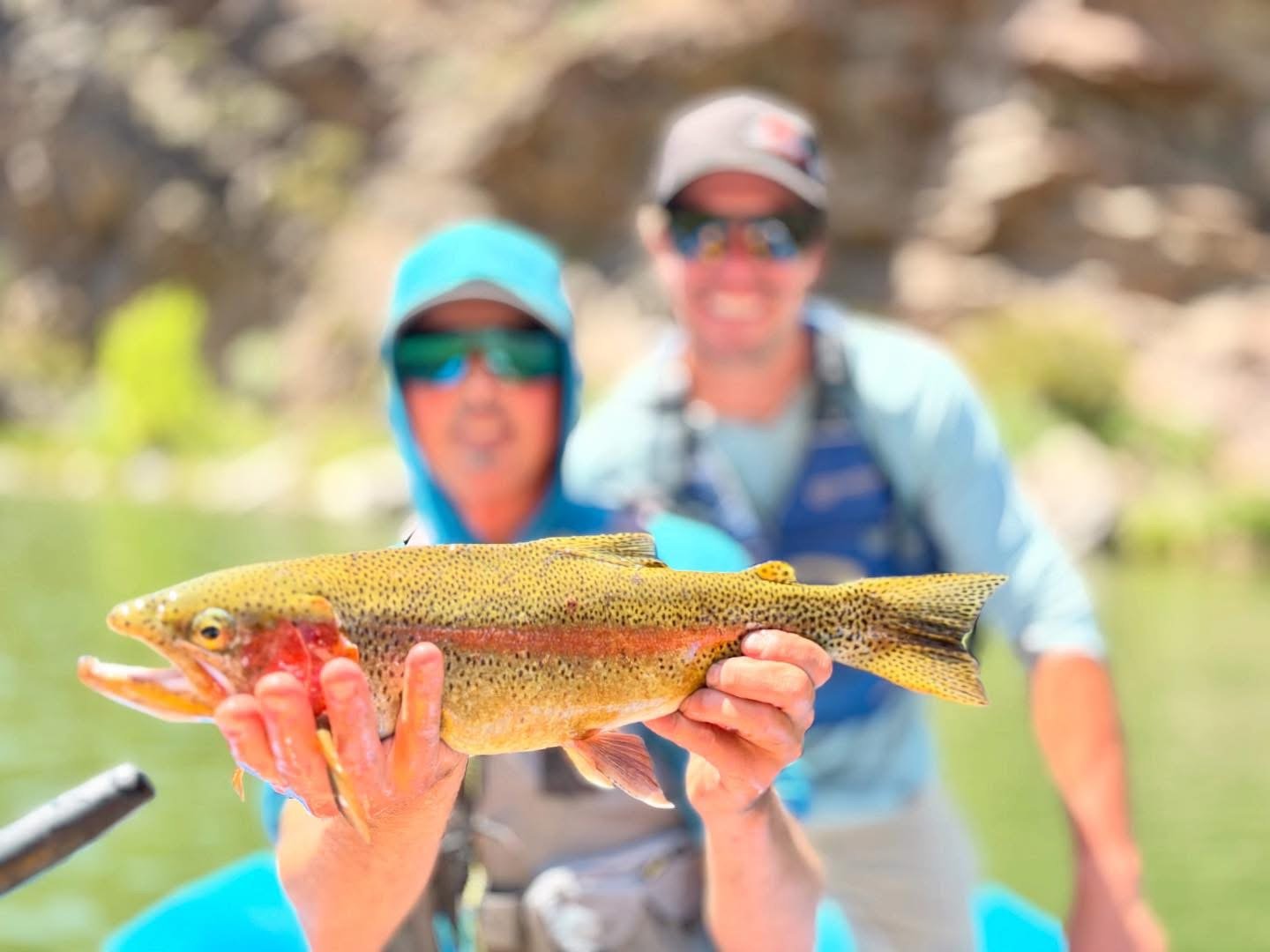 Two men on a boat in a river holding a rainbow trout caught while fishing.