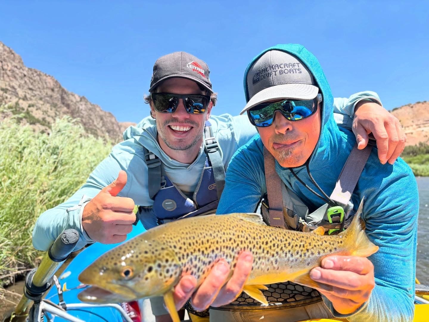 Two men outdoors holding a rainbow trout fish and smiling, wearing sunglasses and outdoor gear, with a mountain and blue sky background.