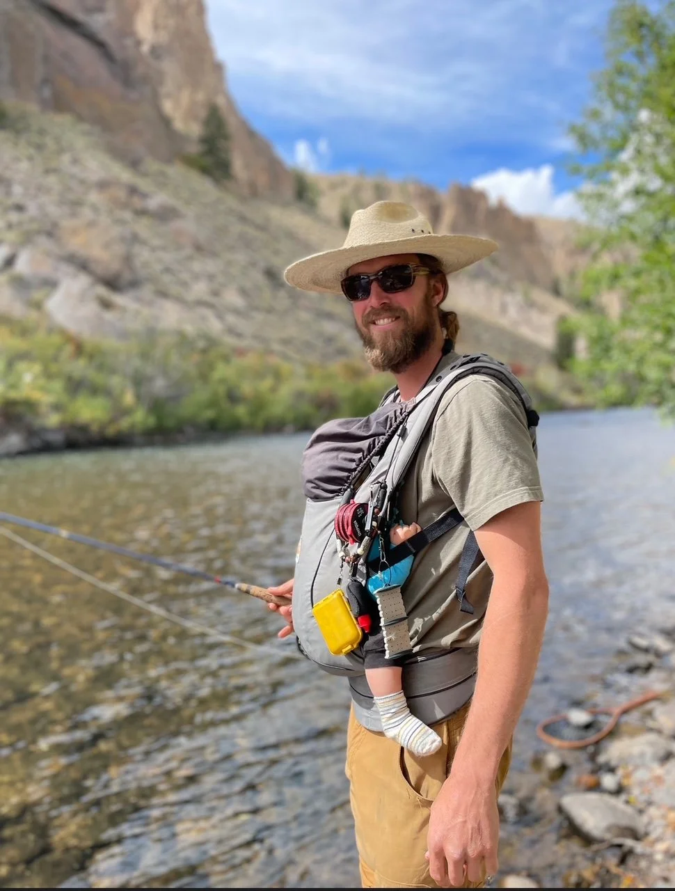 Man fishing in a river while wearing a wide-brimmed hat, sunglasses, a gray t-shirt, khaki shorts, and a backpack with various gear, with mountains and blue sky in the background.