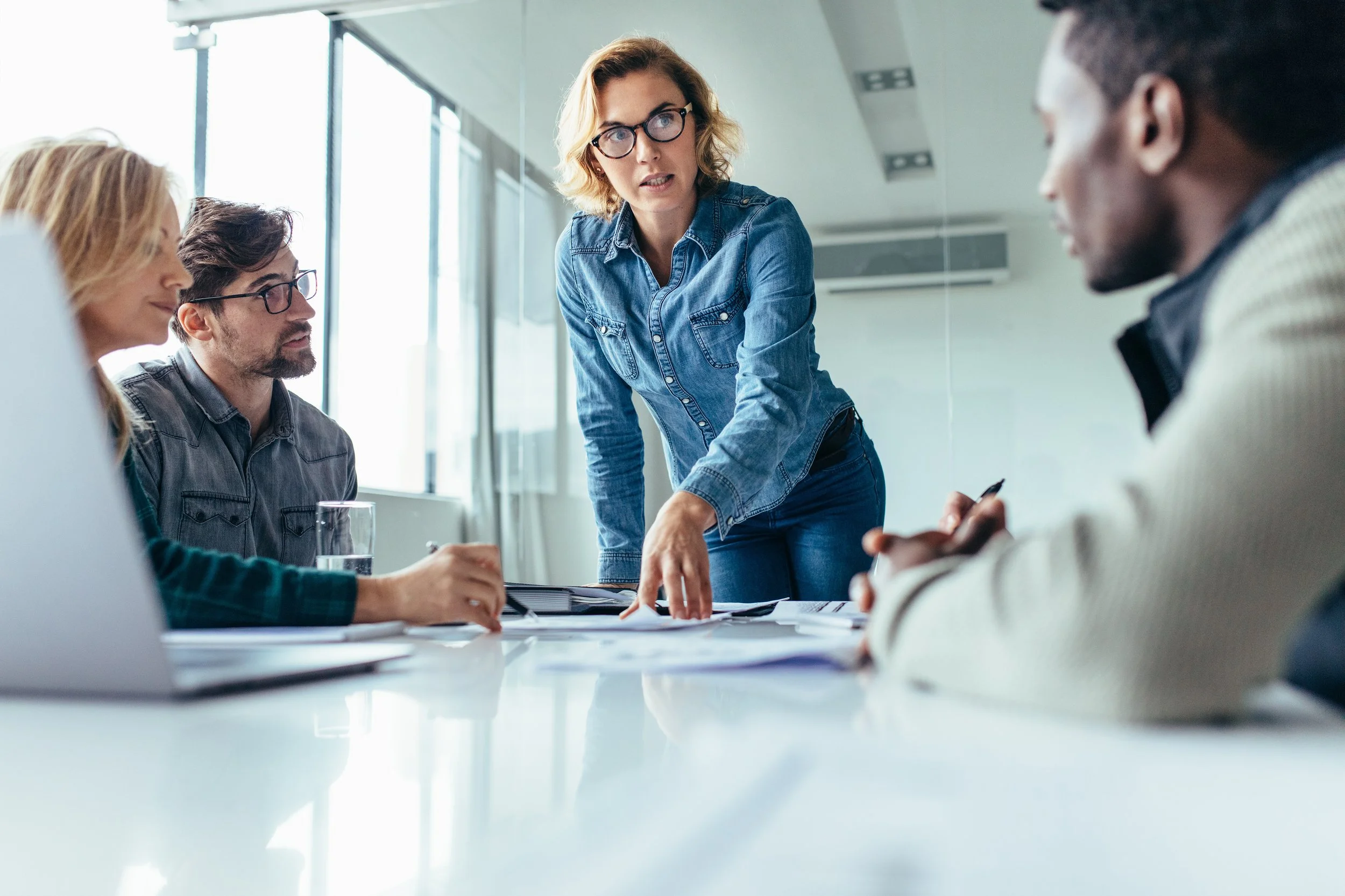 Business meeting with a woman standing and speaking to three seated colleagues in a bright, modern conference room.