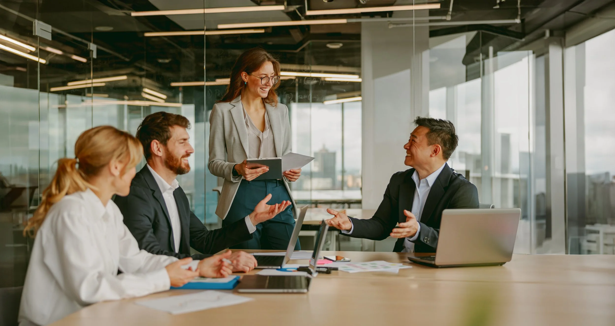 A group of four diverse business professionals in a modern office, engaged in a discussion. One woman is standing, holding a notebook and smiling, while three seated colleagues are listening and responding. The office has large windows, desks with laptops, and documents.
