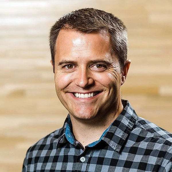 A smiling man in a blue and black checkered shirt standing in front of a wooden wall.