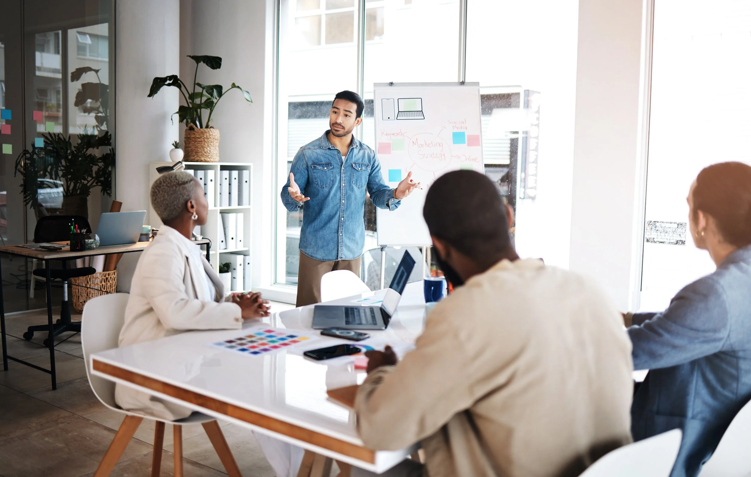 A man in a blue denim shirt is presenting to a group of four people in a modern office, standing in front of a whiteboard with notes on marketing strategy, while the others sit around a table with laptops and documents.