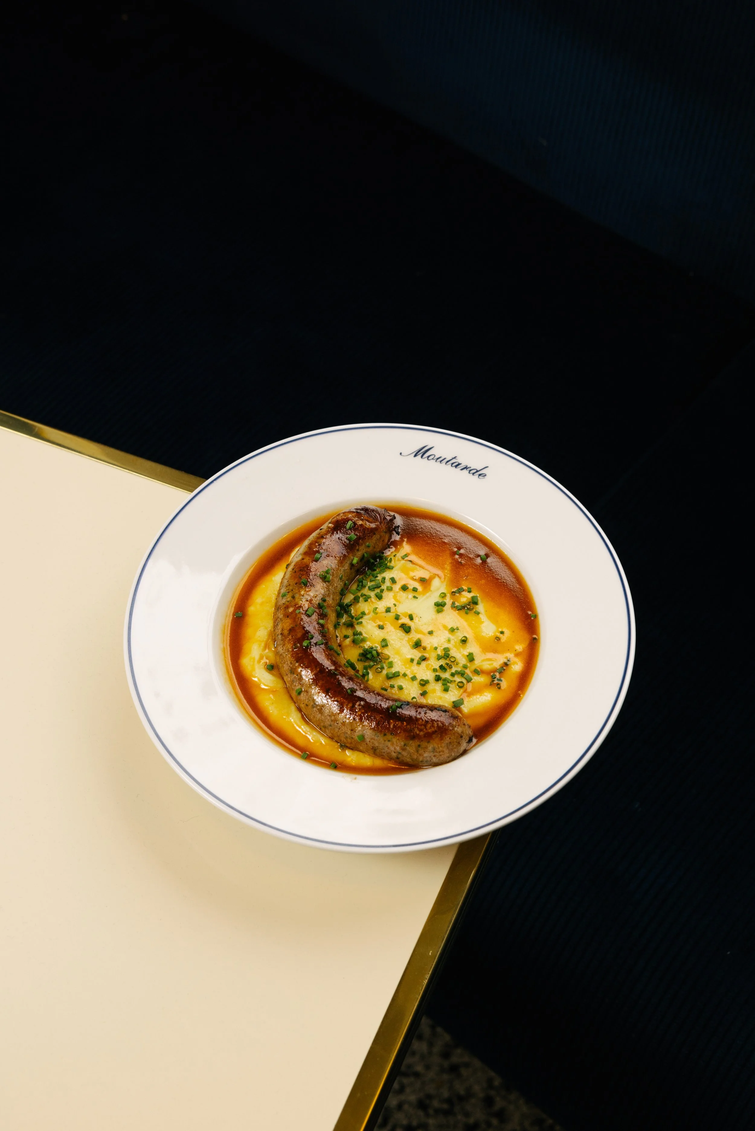 Assiette de purée de pommes de terre avec une saucisse, garnie de moutarde et de ciboulette, sur une table de restaurant.