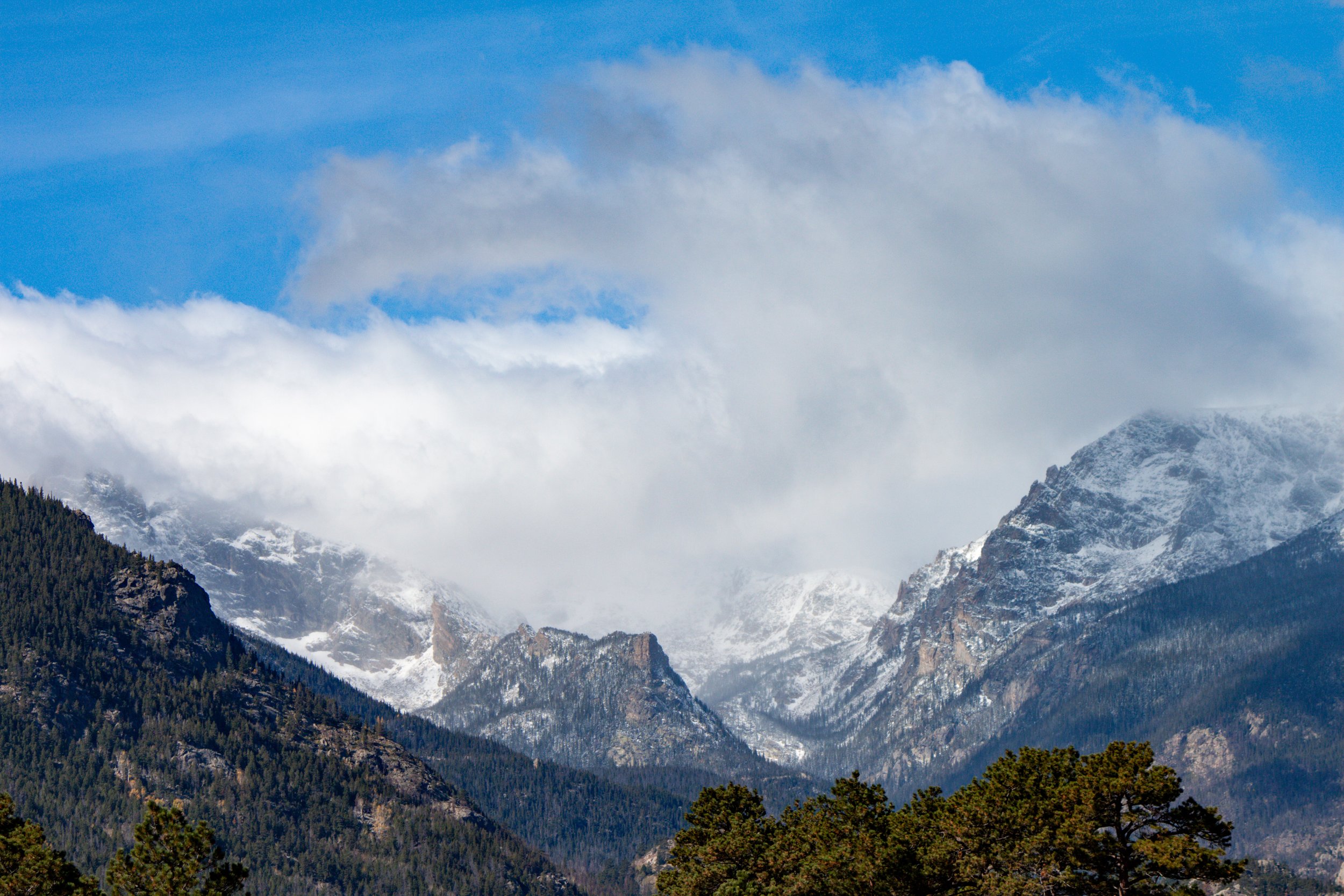 Snow-capped mountains partially covered by clouds with trees in the foreground and a blue sky above.