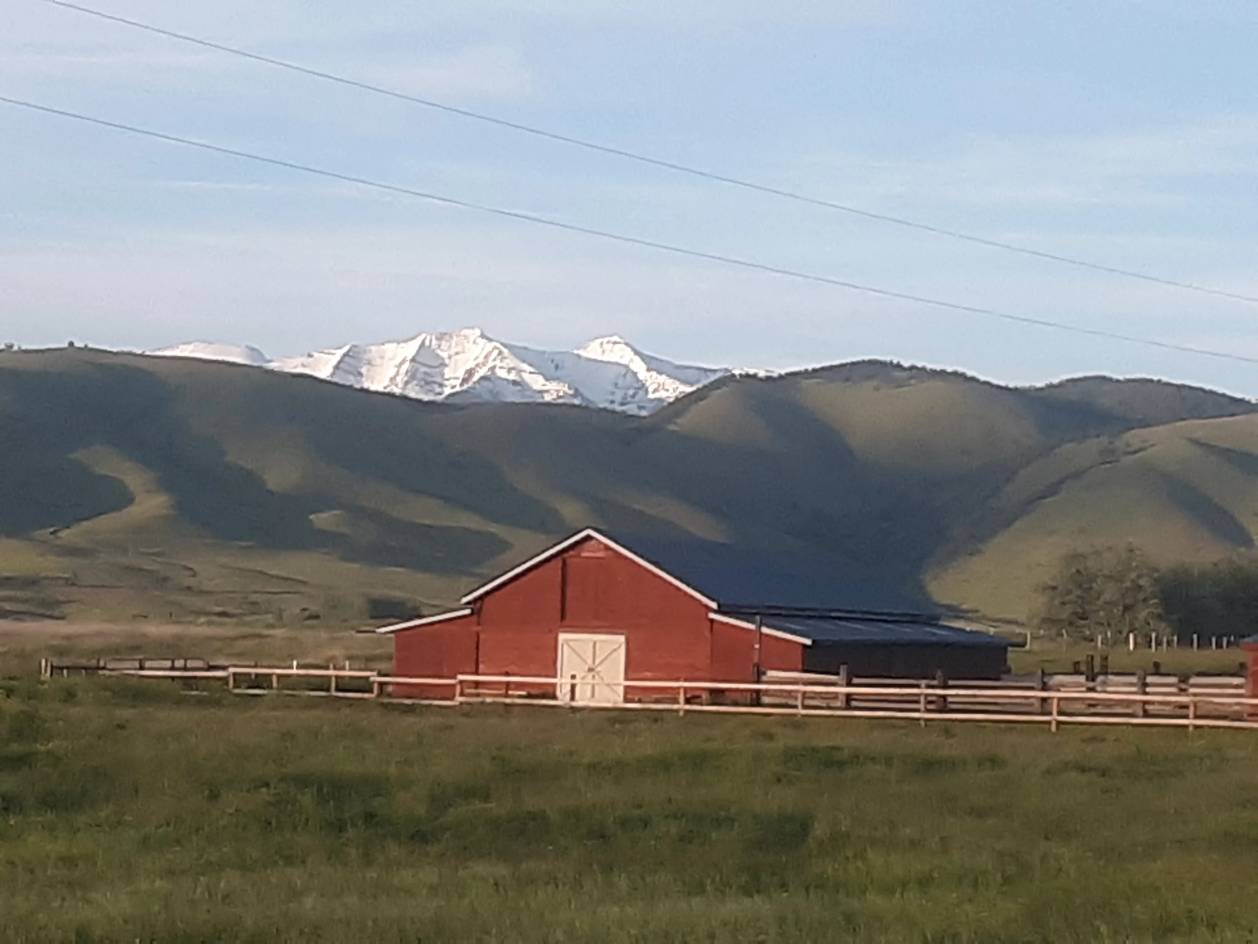 Red barn with white doors in a green field, hills in the background, snow-capped mountains further in the distance, and a blue sky with some clouds.
