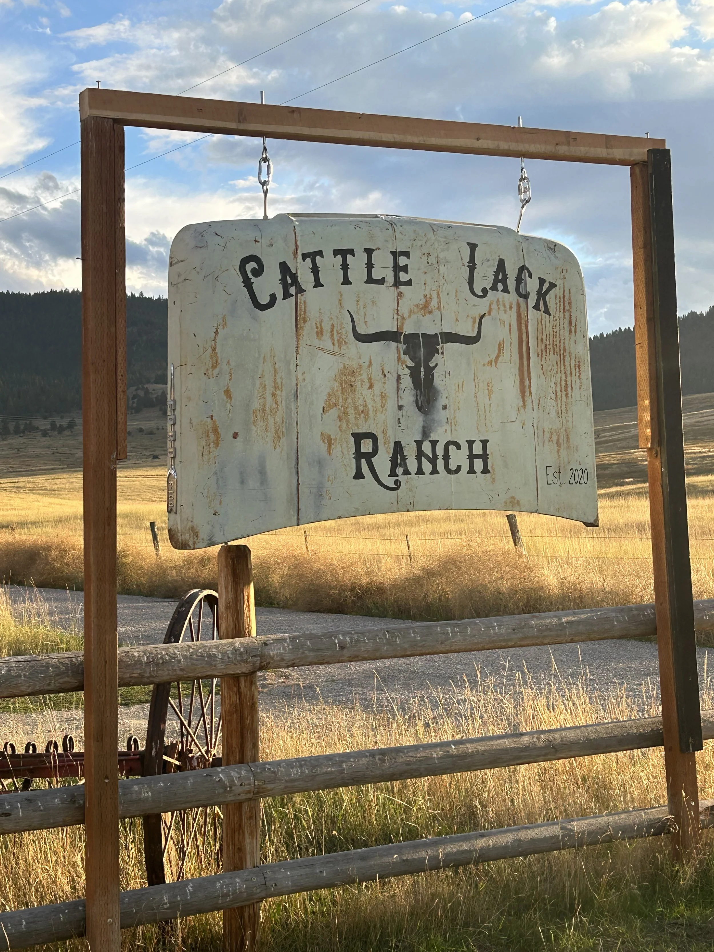 A rustic sign for Cattle Lack Ranch, featuring a bull skull with horns, hanging from a wooden frame, set in a rural landscape with fields and mountains in the background.