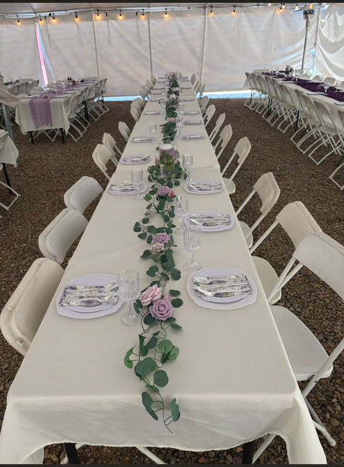 Long rectangular banquet table with white tablecloth and foldable chairs, decorated with a green garland and purple roses in the center, set for a meal with plates, napkins, and glassware, under a tent with string lights.