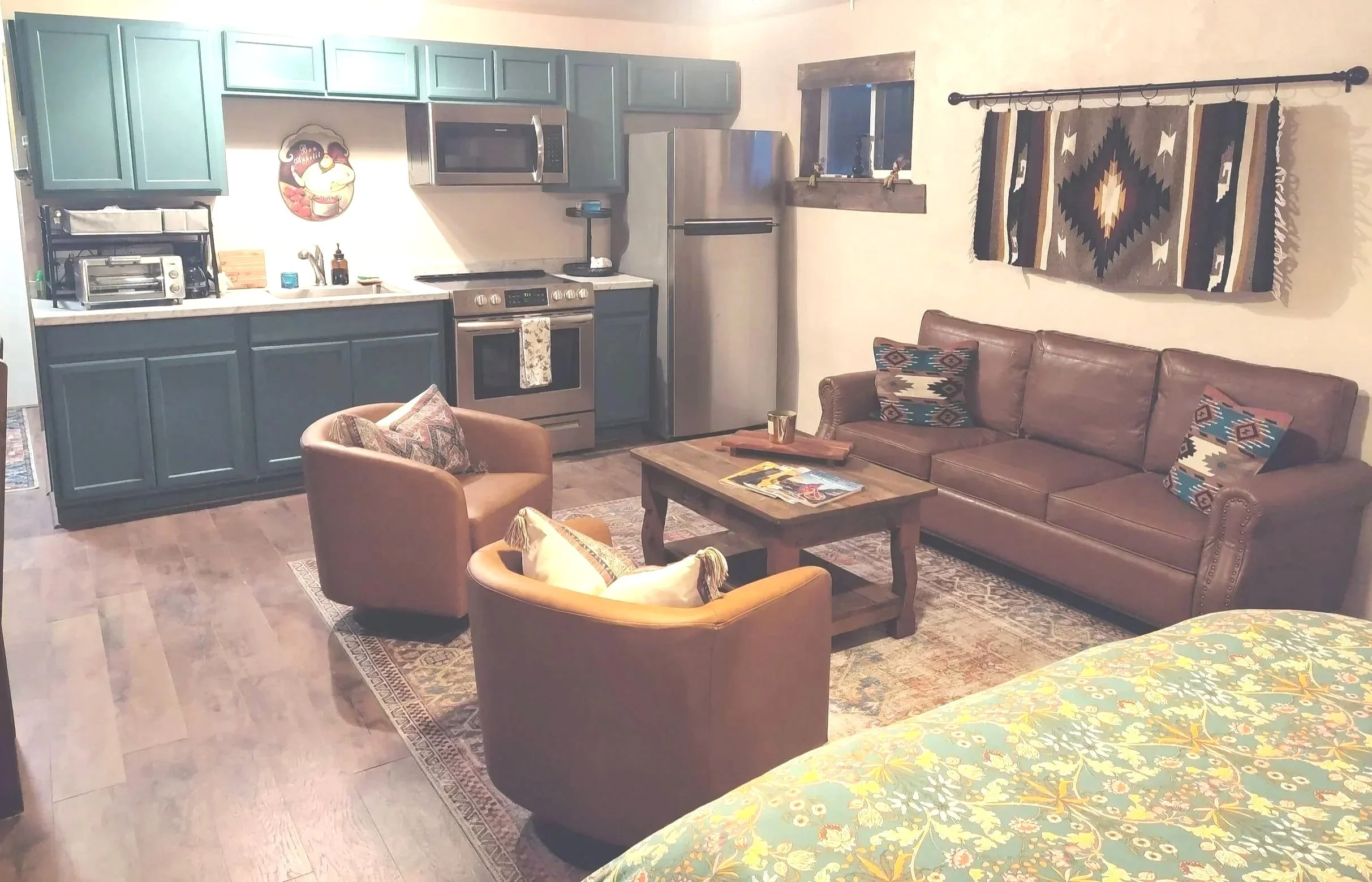 cozy living room with a kitchen in the background. There is a brown leather sofa with patterned pillows, a coffee table with magazines and a mug, and an armchair with a pillow. The kitchen has green cabinets, a stainless steel refrigerator, microwave, and oven. A tapestry hangs on the wall above the sofa, and a window with a wooden frame is visible.