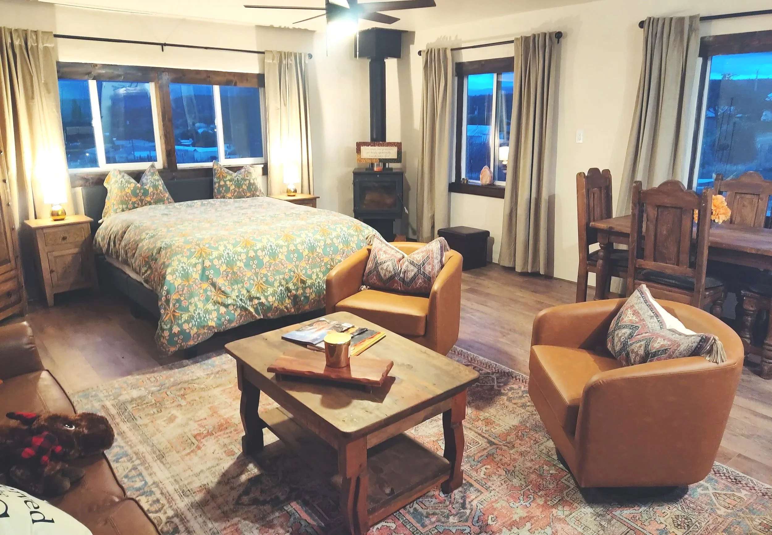 An intimate adobe studio casita in a village along the High Road to Taos featuring a cozy living room with a bed near a window, a wooden table, armchairs, and a fireplace, with curtains covering the windows and a ceiling fan overhead.