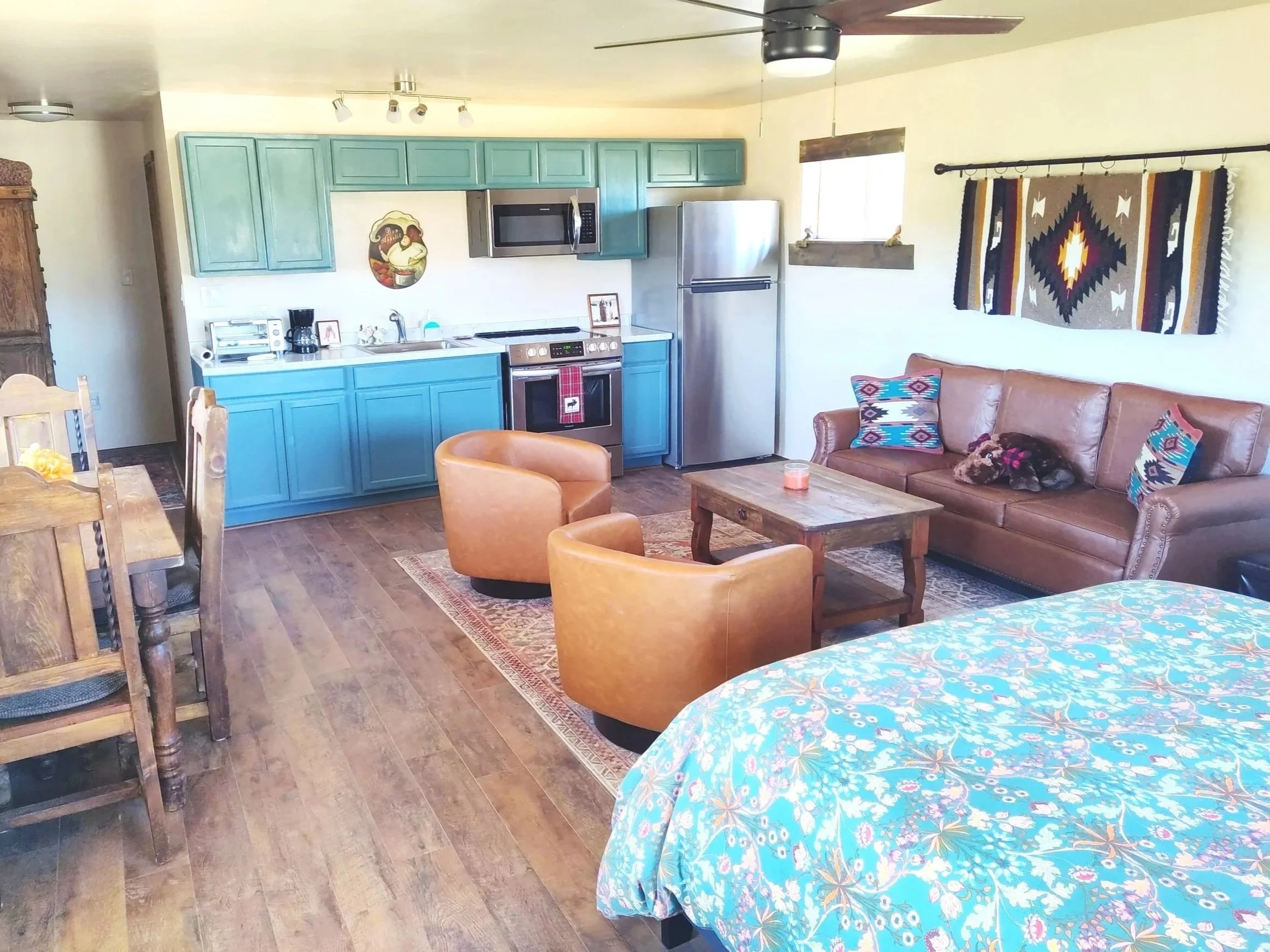A cozy living room with blue kitchen cabinets, a stainless steel fridge, a brown leather sofa with colorful patterned pillows, and wooden furniture. There is a woven wall hanging above the sofa and a small coffee table with a candle. The room has wooden flooring and a window letting in natural light.