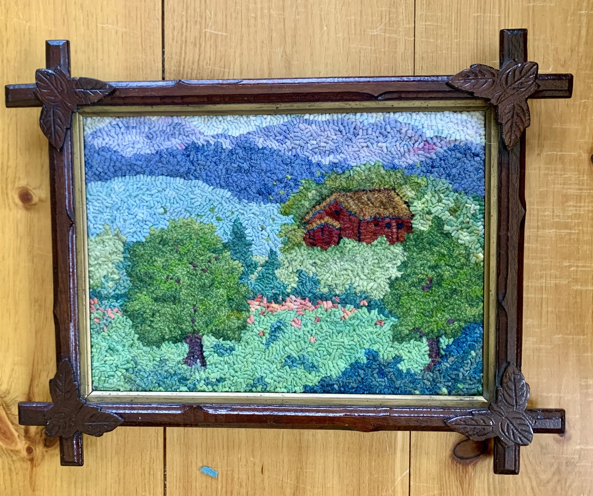 Colorful embroidered landscape showing trees, a house, mountains, and a sky with clouds in a wooden frame with leaf carvings.