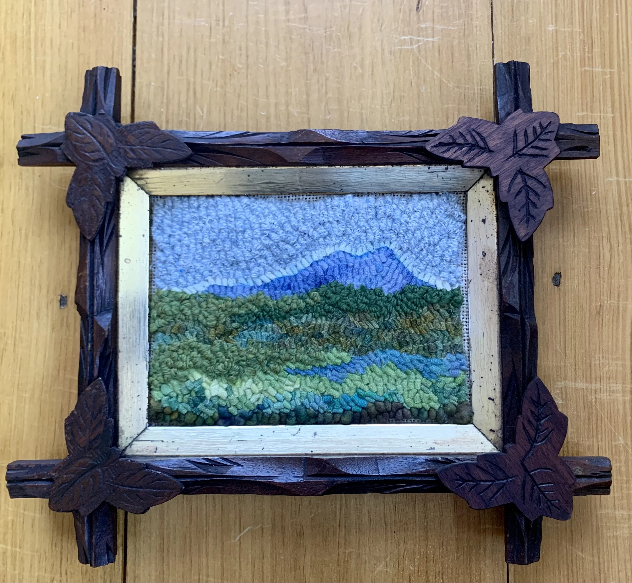 Framed landscape embroidery of mountain, sky, and green hills with a wooden leaf corner frame.