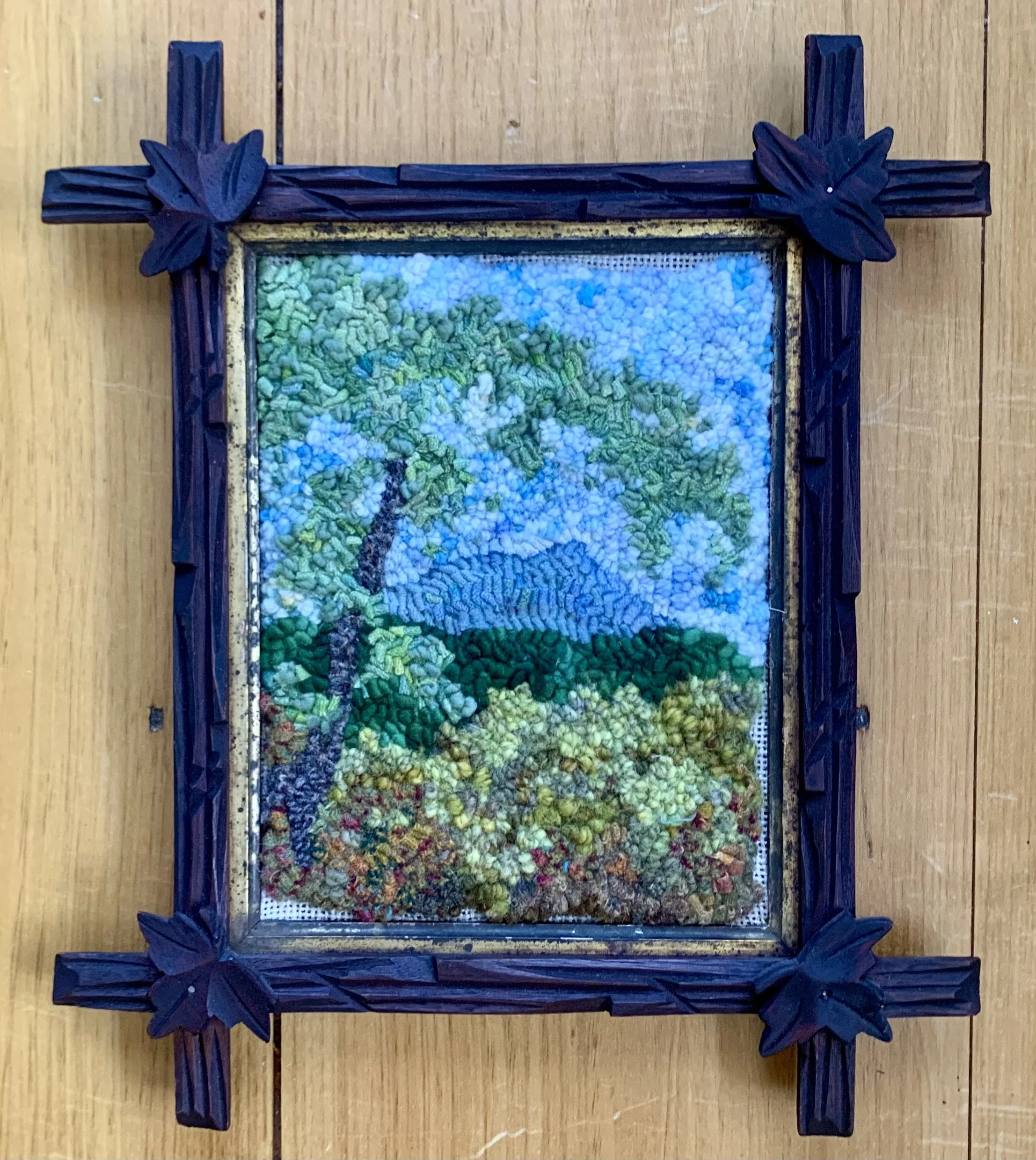 Framed cross-stitch artwork of a landscape with a tree, hills, and a blue sky, on a wooden surface.