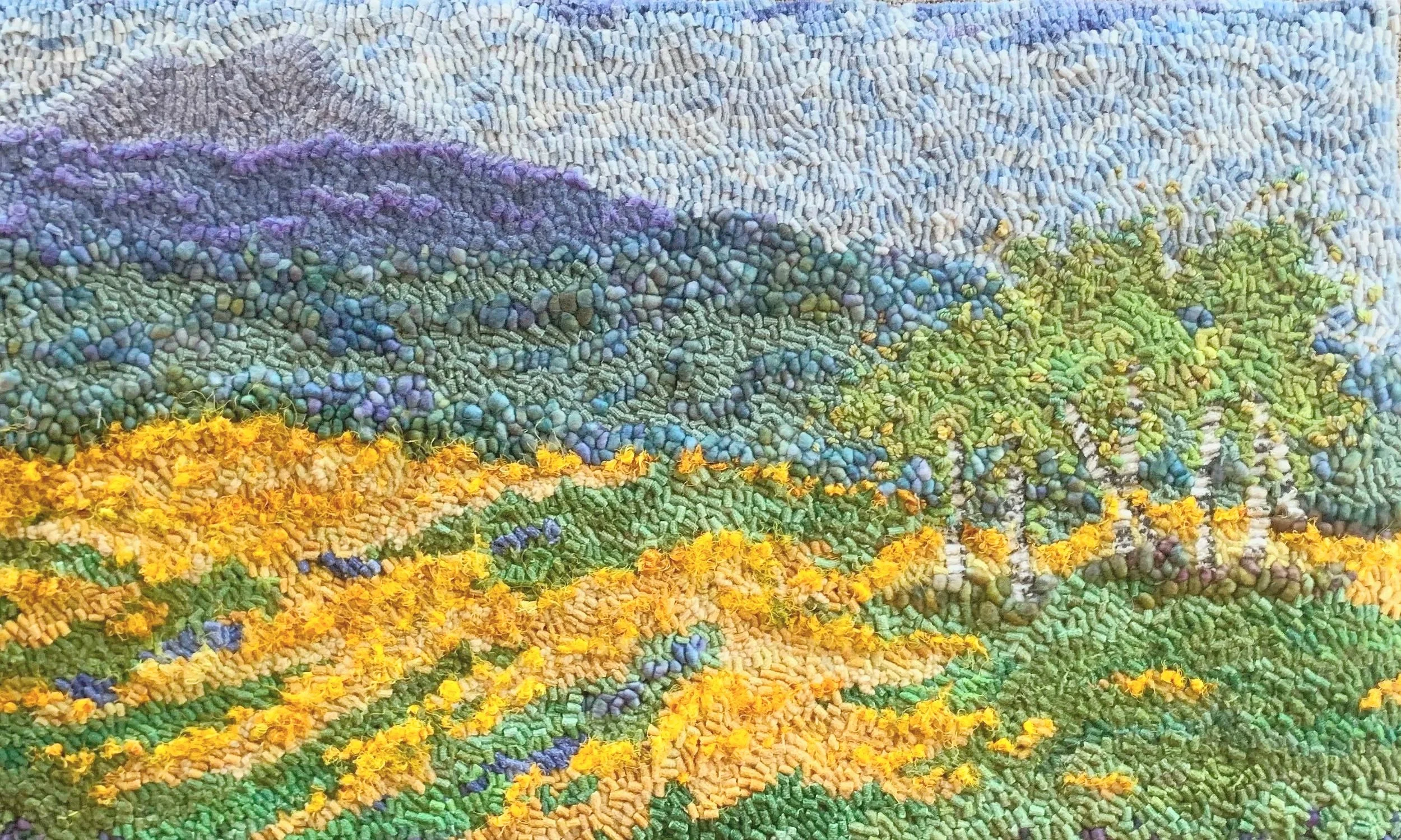 Textured embroidery depicting a vibrant landscape with rolling hills, flowering fields in yellow and blue, densely treed green areas, and purple mountains in the background.