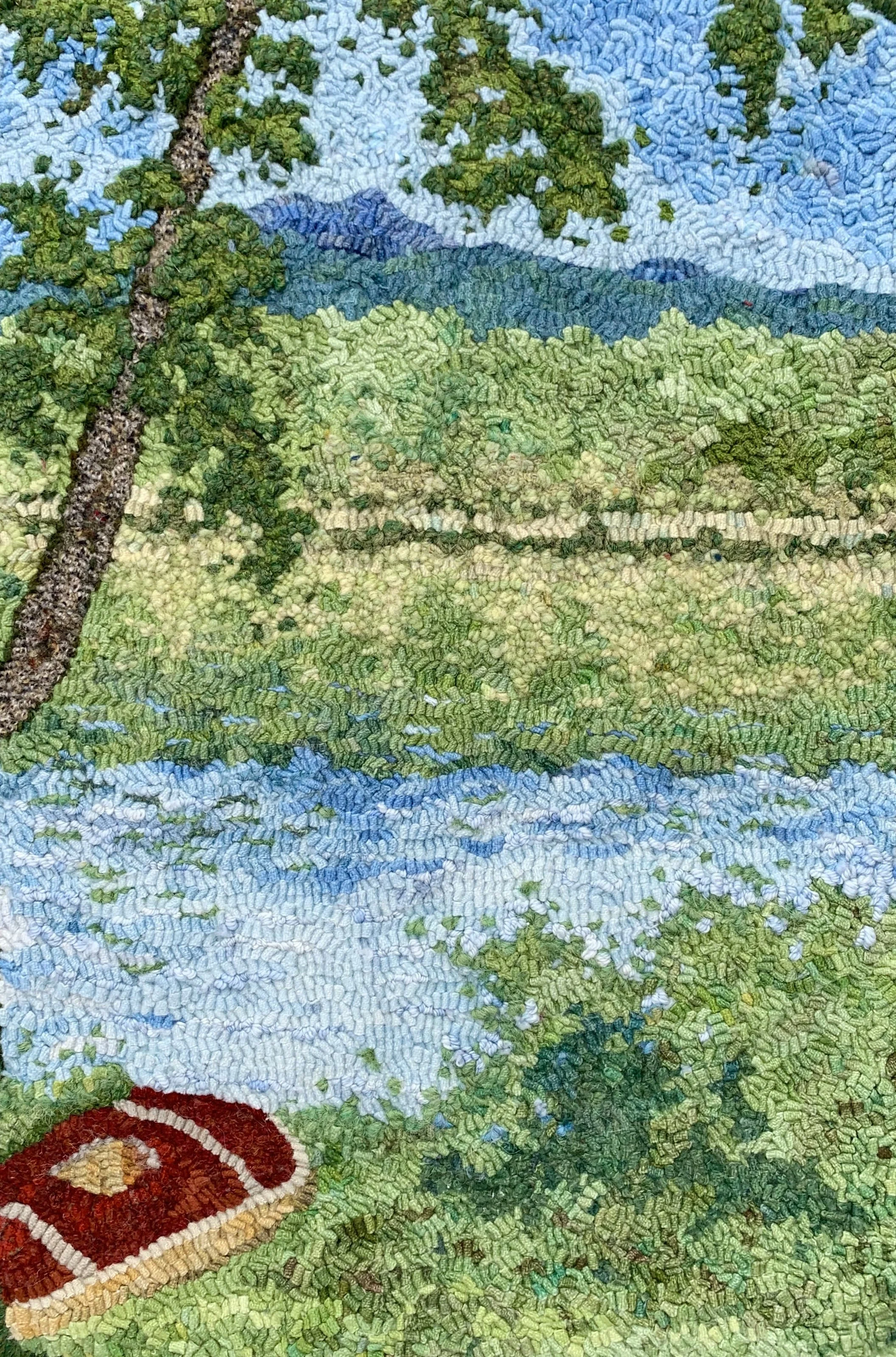 Close-up of a tapestry or embroidery depicting a landscape scene, including a tree, green hills, a blue lake or river, and mountains in the background with blue sky.