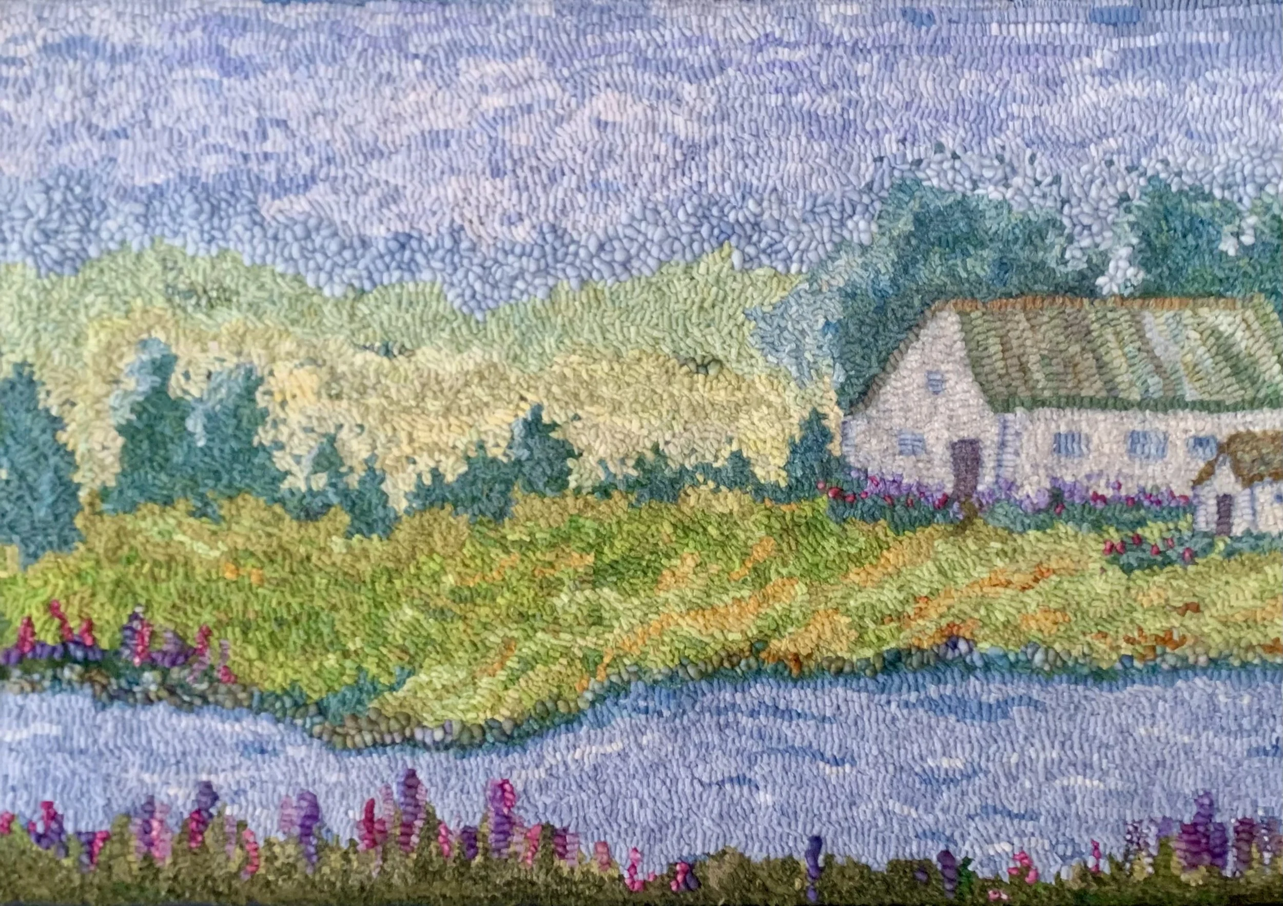An embroidered landscape scene featuring rolling hills, a small white house with a green roof, trees, and vibrant flowers along a water body.
