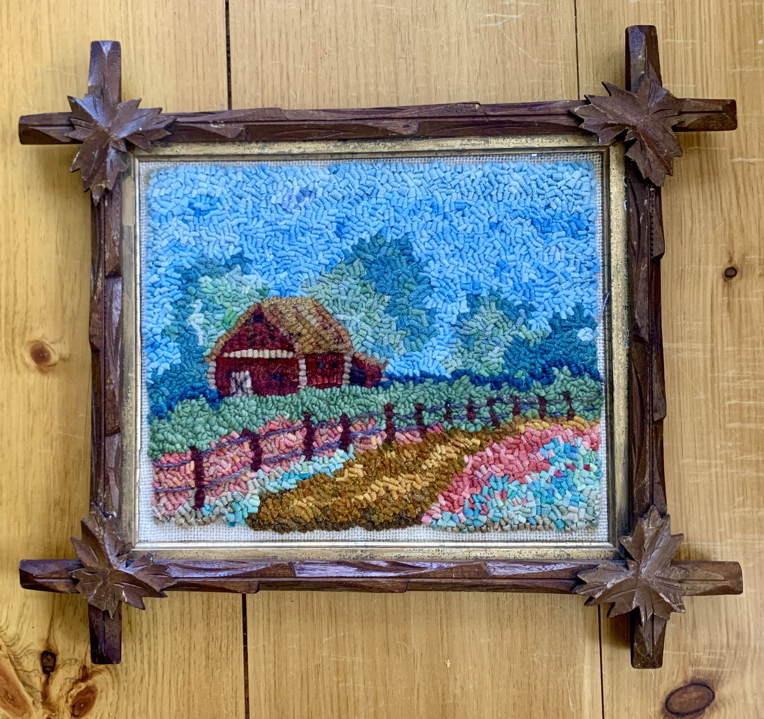 Colorful pixel art embroidery of a countryside scene with a barn, trees, a fence, and mountains, framed with carved wooden leaves.