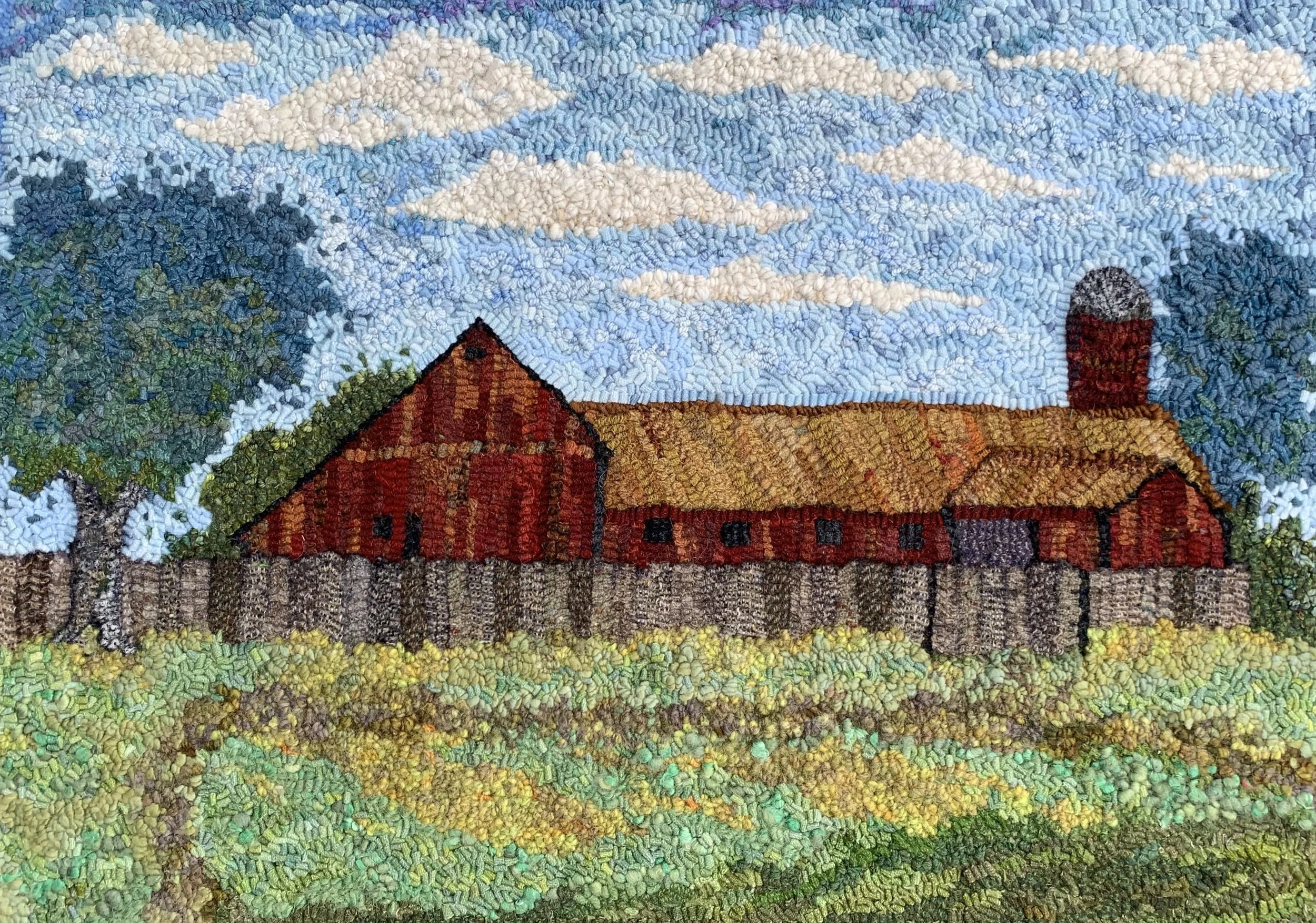 A textured embroidery art depicting a rural farm scene with a large red barn, a silo, trees, a wooden fence, and a cloudy blue sky.