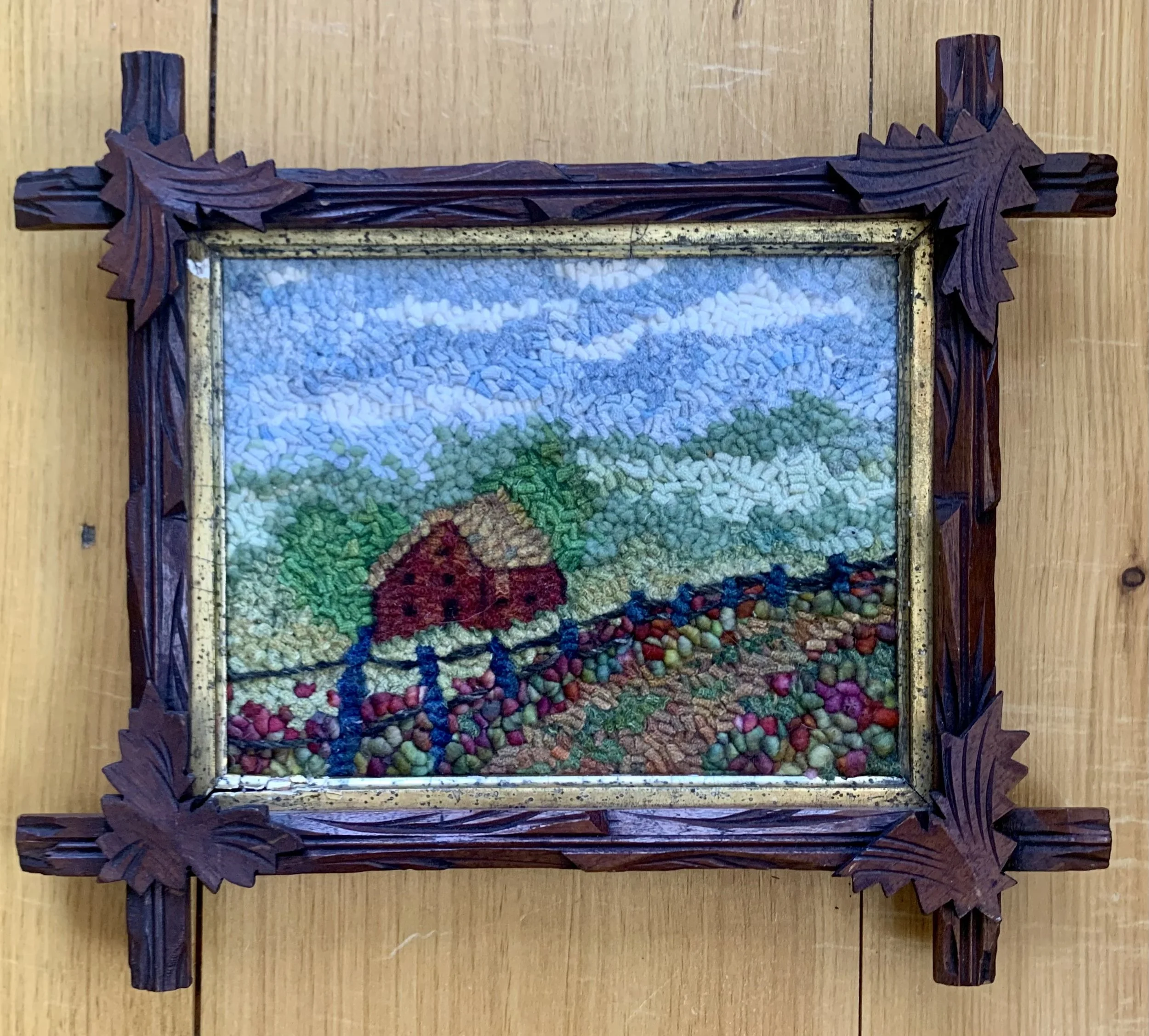 A framed mosaic artwork depicting a rural landscape with a red barn, green trees, hills, and a blue sky, mounted on a wooden wall.