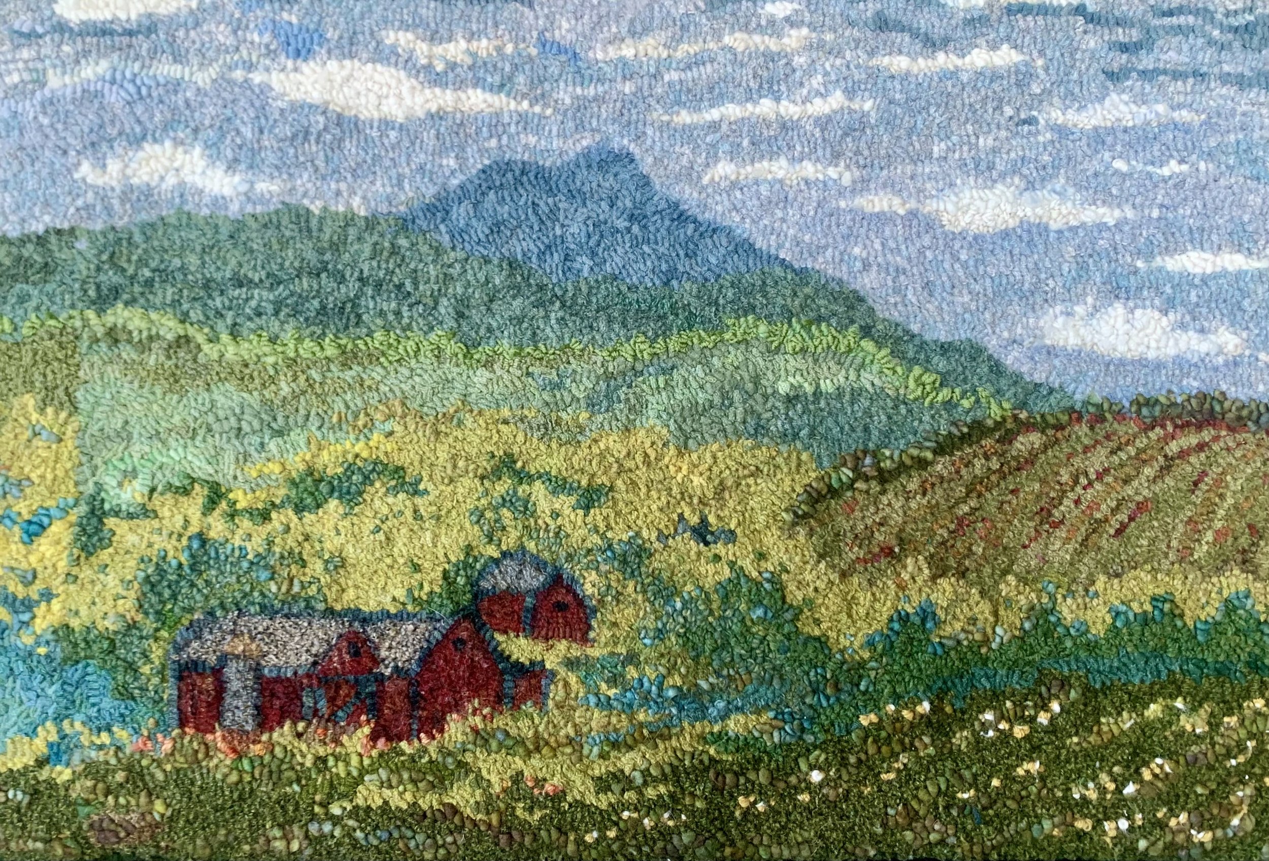 A detailed embroidered landscape featuring rolling green hills, yellow fields, and a blue mountain in the background, with a bright sky and scattered clouds.