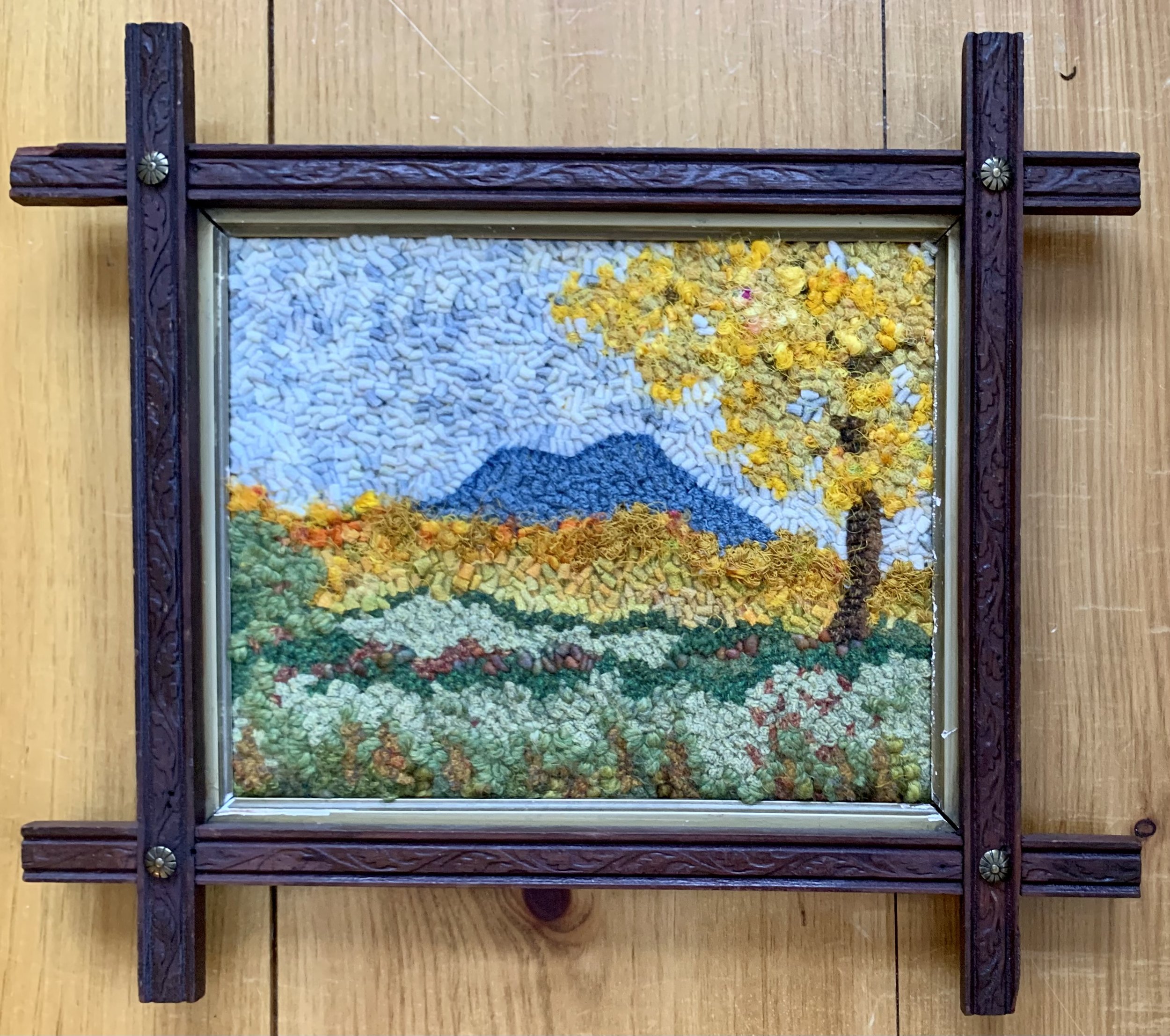 A framed landscape scene of a mountain, yellow trees, and green fields, made with textured, colorful embroidery or yarn on fabric.
