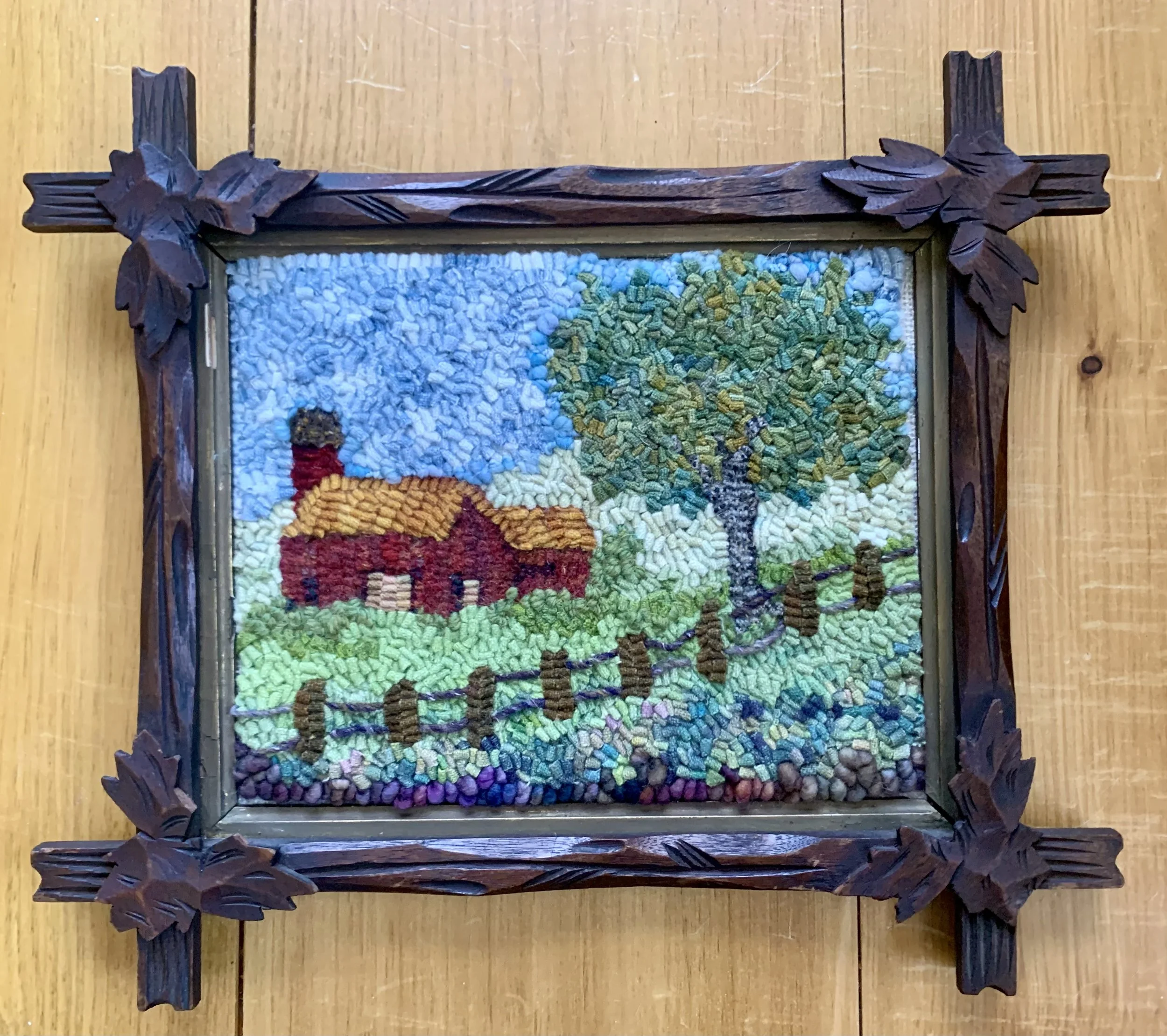 Framed artwork depicting a landscape scene with a red house, chimney smoke, large tree, and grassy area under blue sky.