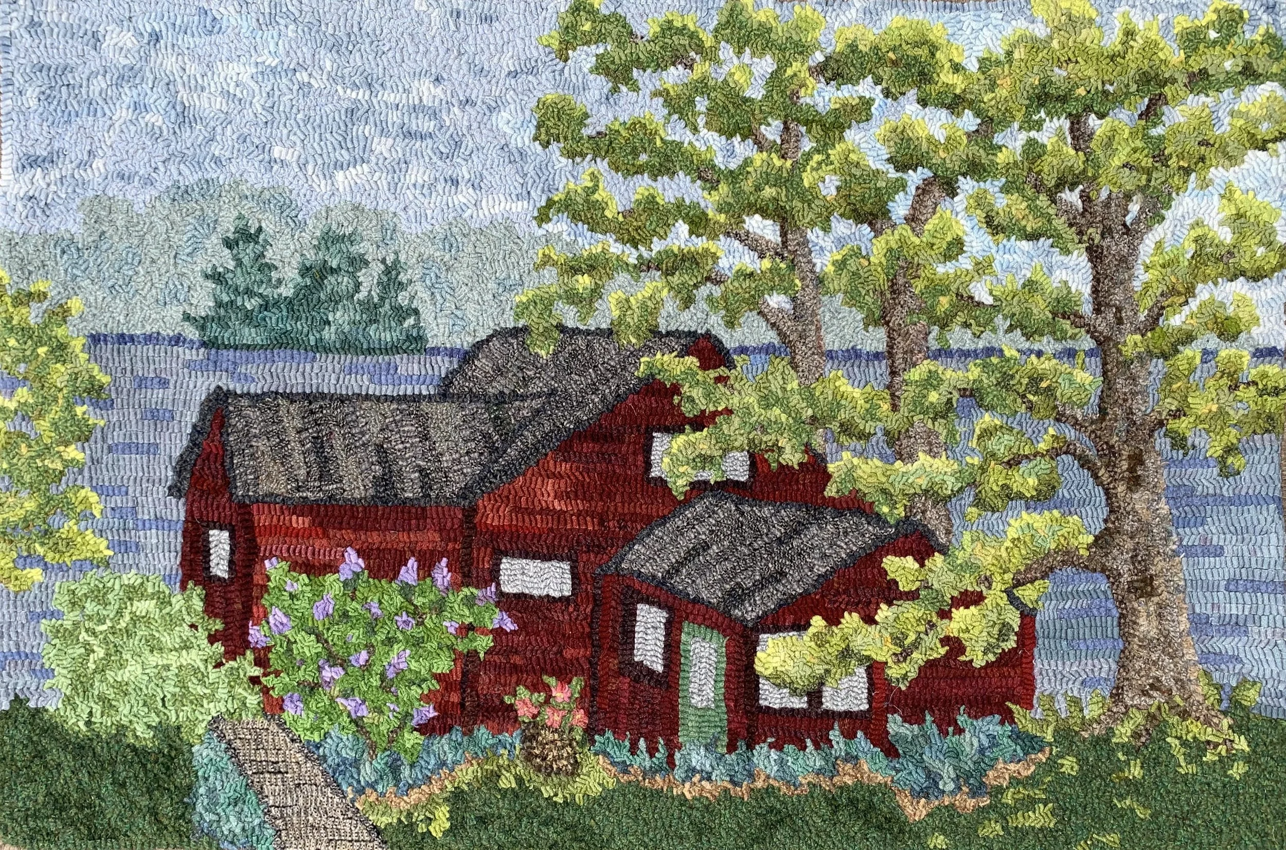 A detailed embroidered picture of a red house surrounded by green trees and plants, with a pond in the background and a blue sky overhead.
