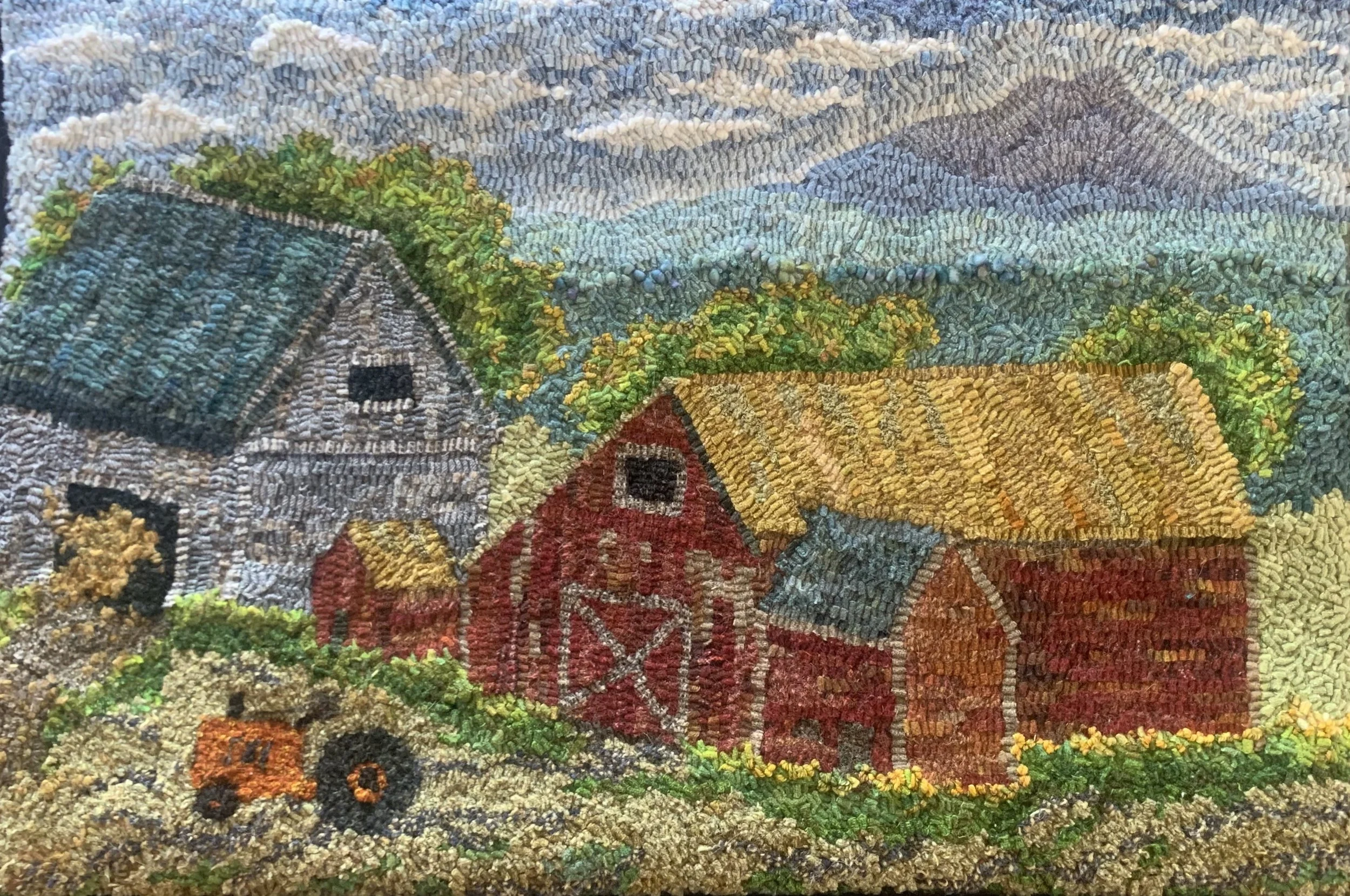 A colorful rustic farm scene created with embroidery, featuring a barn with a yellow roof, a red barn with a gray roof, a gray house with a green roof, a small red and orange tractor, trees, bushes, and a sky with clouds and mountains in the background.