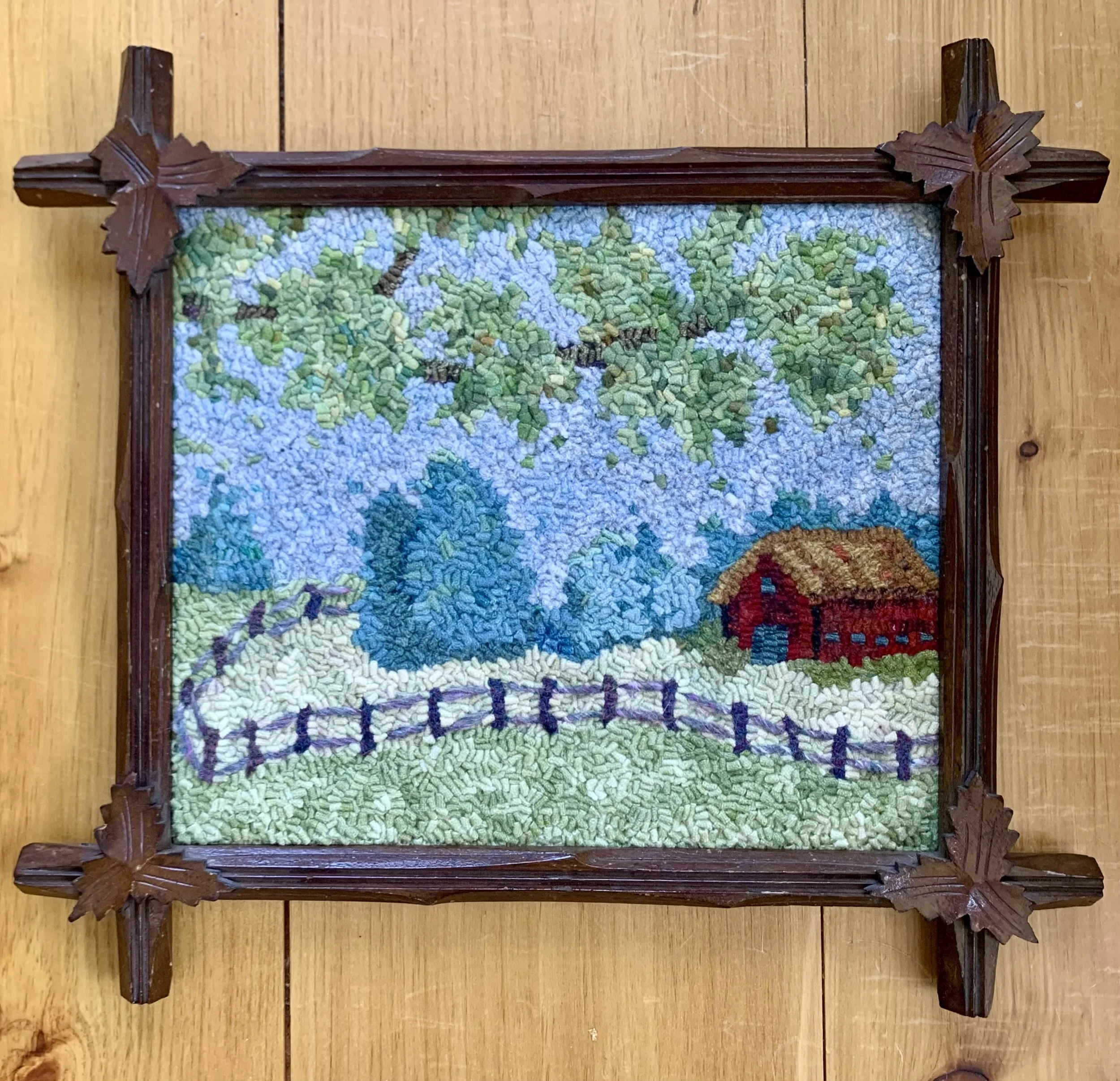 A framed embroidery artwork depicting a rural farm scene with a red barn, green trees, a white fence, and a blue sky, mounted on a wooden surface.