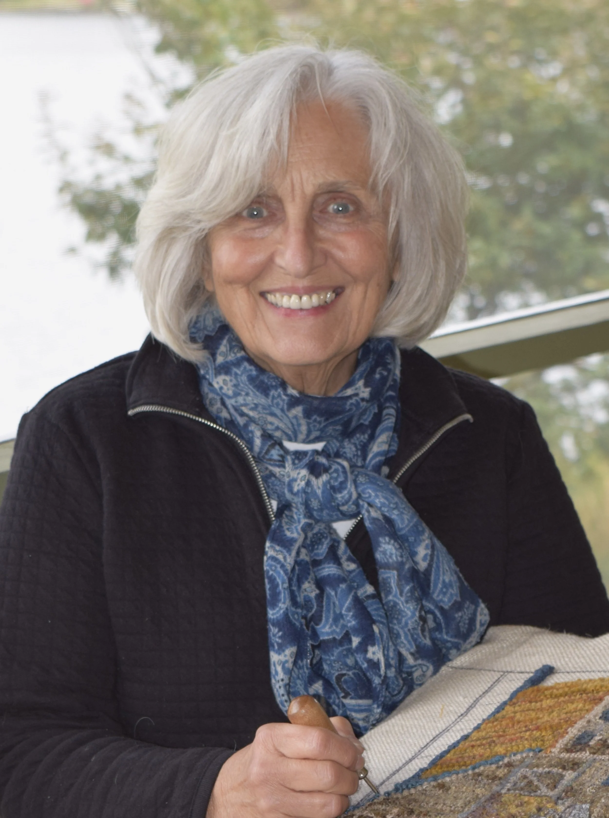An elderly woman with gray hair and blue eyes, smiling, holding a needle and working on a colorful quilt.