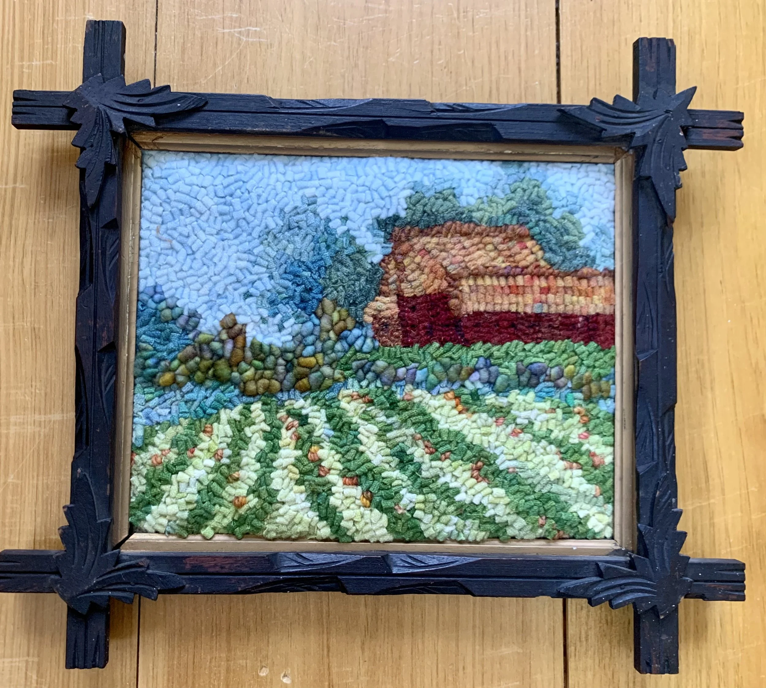 A framed artwork made of colorful yarn depicting a rural farm scene with a barn, green fields, cultivated land, trees, and a blue sky.