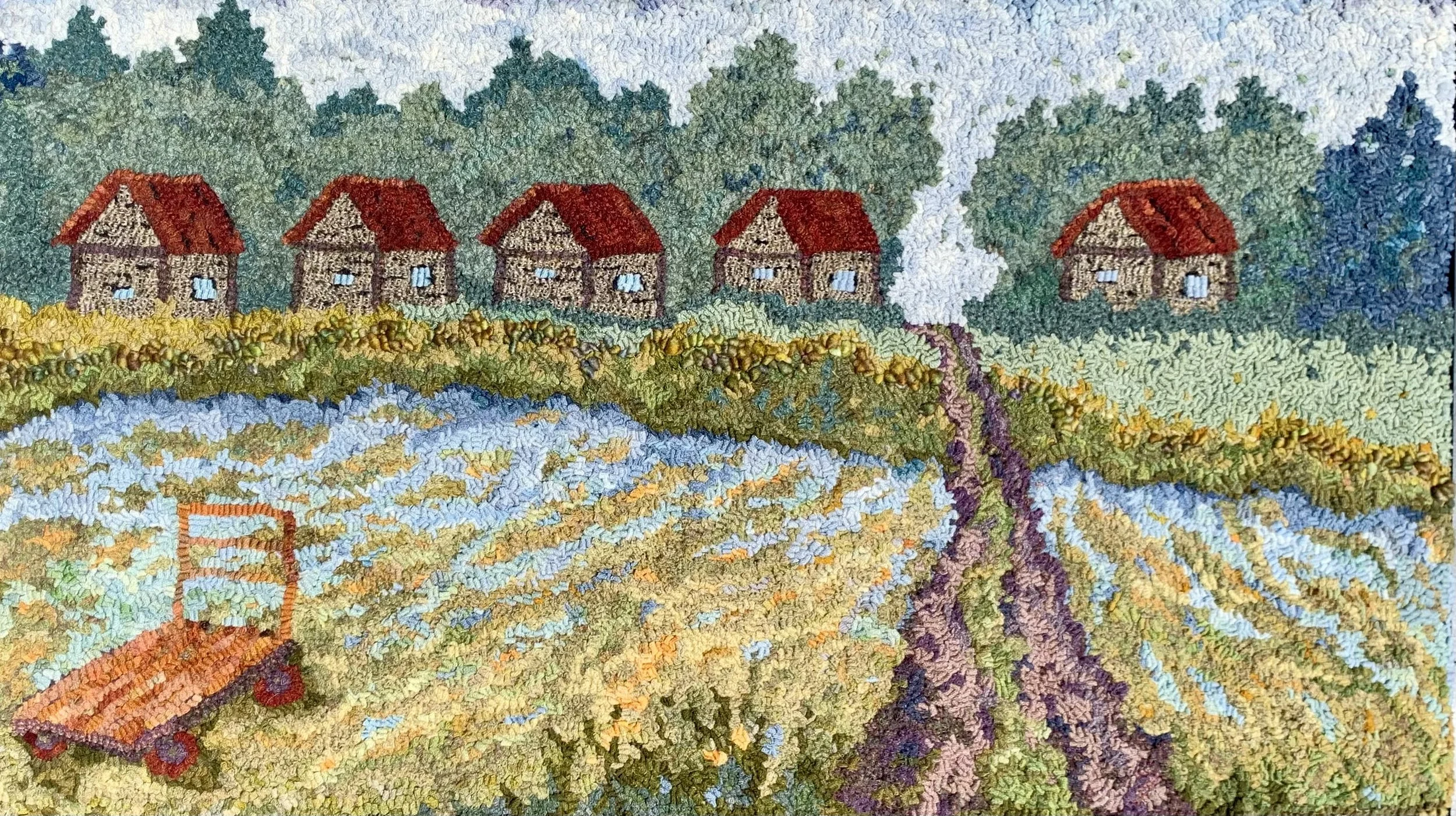 A colorful embroidered landscape depicting five houses with red roofs, trees, a river, fields, and a cart on a grassy path.