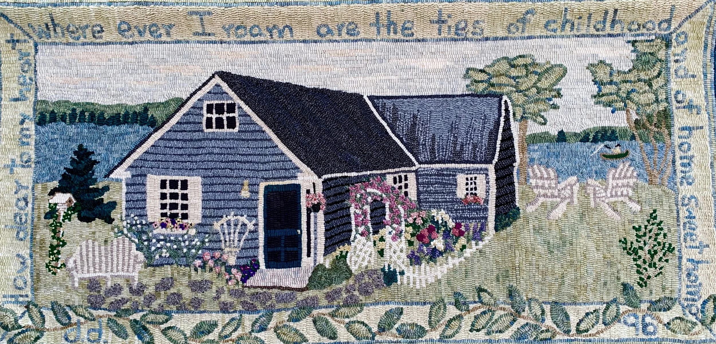 A detailed embroidery of a blue house with flower gardens, trees, a lake with a boat, and a bench, surrounded by a decorative border with text at the top.