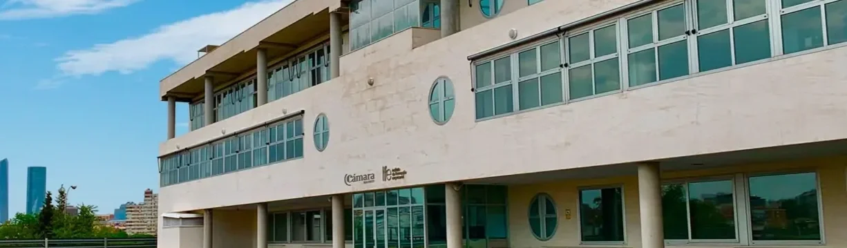 Edificio moderno con paredes de color arena y amplias ventanas de vidrio con marco metálico, en un entorno urbano con cielo azul y algunas nubes.