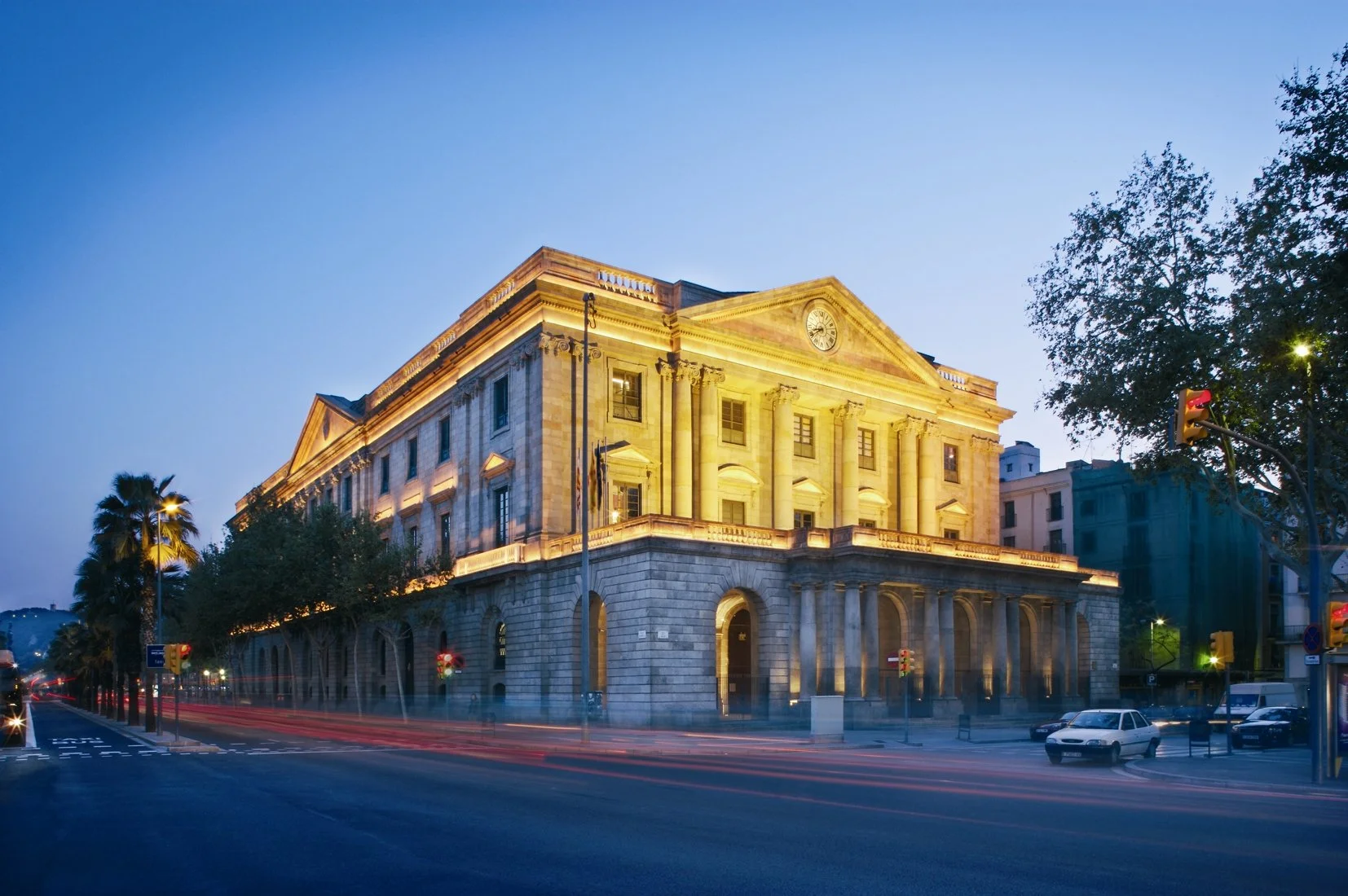 Edificio histórico iluminado en la noche, con columnas en la entrada y un reloj en la cima, en una esquina de calle con árboles y semáforos.