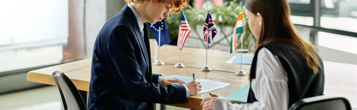 Dos personas en una reunión formal en una oficina, firmando documentos. Sobre la mesa hay banderas de diferentes países.
