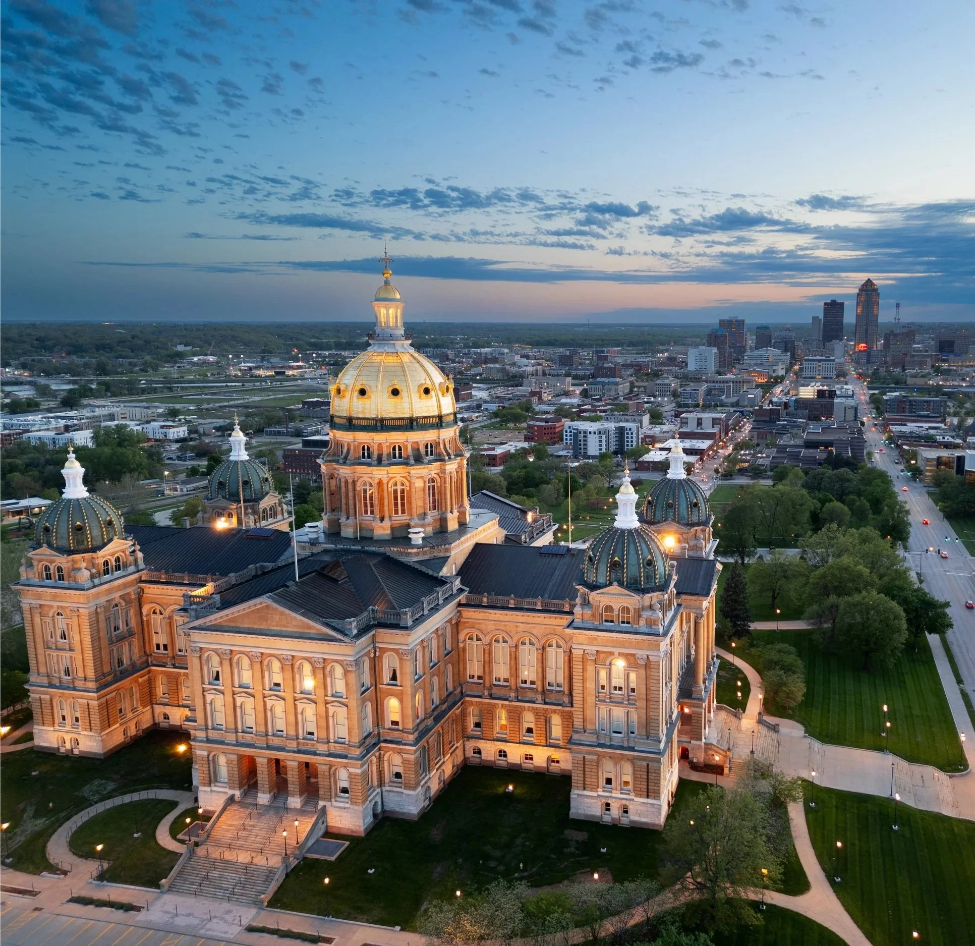 Vista aérea de un majestuoso edificio con cúpulas y luminarias en la ciudad durante el atardecer, con cielo parcialmente nublado y calles iluminadas.