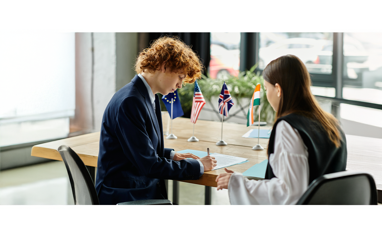Dos personas firmando documentos en una mesa de oficina, con banderas de diferentes países en el fondo.