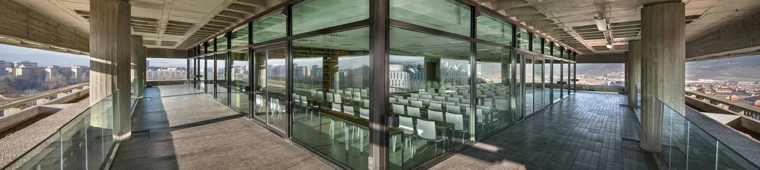 Terraza de un edificio con vista panorámica, con paredes de vidrio, muebles de oficina y vista urbana en el fondo.