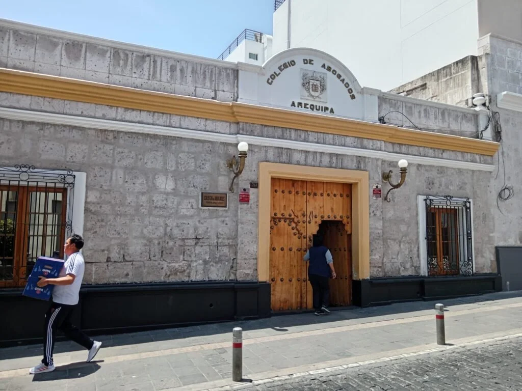 Edificio con un cartel que dice Colegio de Abogados Arequipa, con puertas de madera y ventanas con rejas de hierro, en la calle.