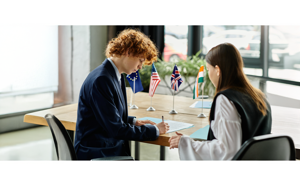 Joven firmando un documento en una mesa con banderas de diferentes países en el fondo, posiblemente en una reunión internacional.