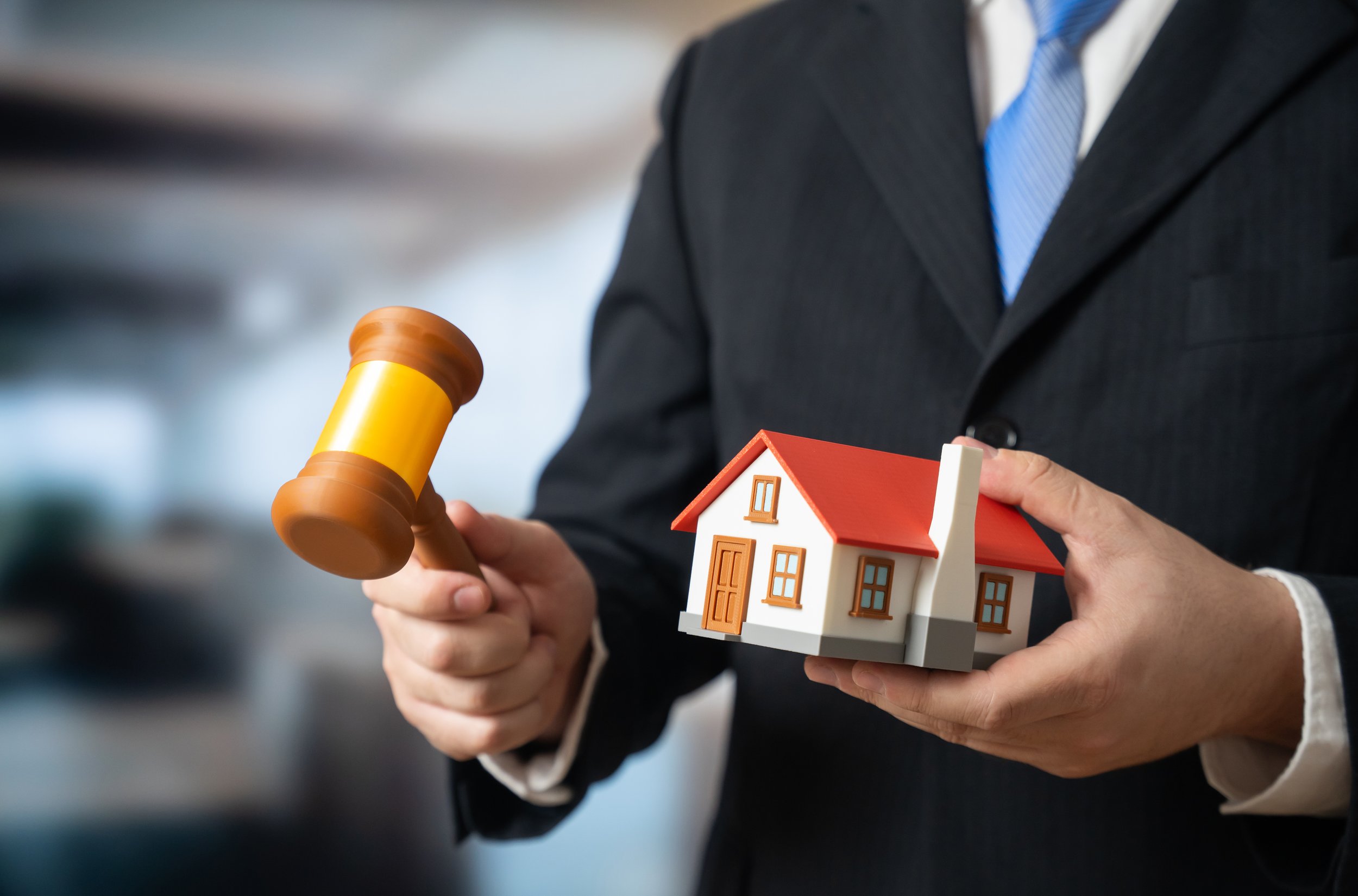 Persona con traje negro sosteniendo un martillo de juez y una maqueta de casa en un fondo difuso.