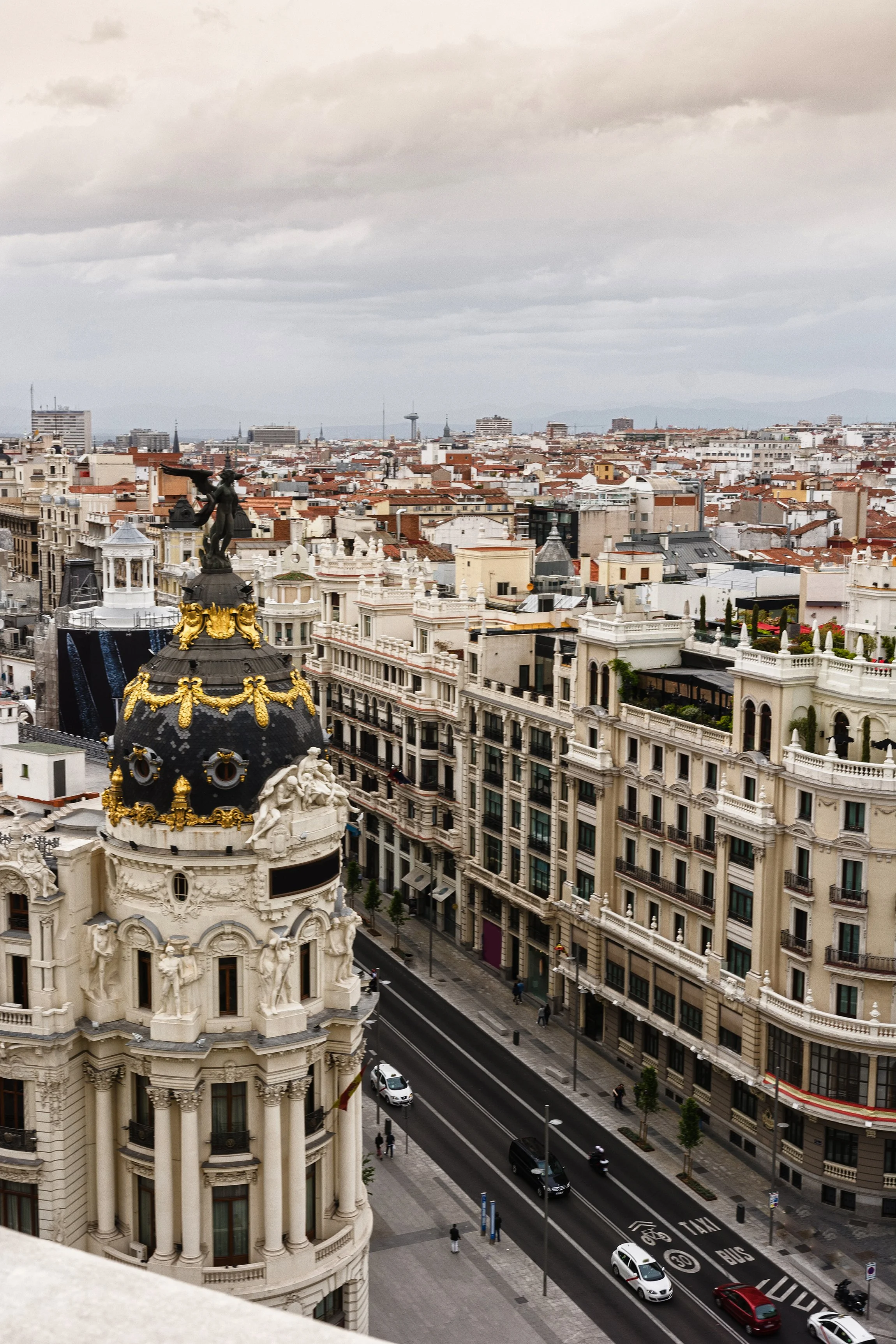 Vista aérea de la ciudad con edificios históricos y modernos, se observa una calle con vehículos y peatones, y un cielo nublado.